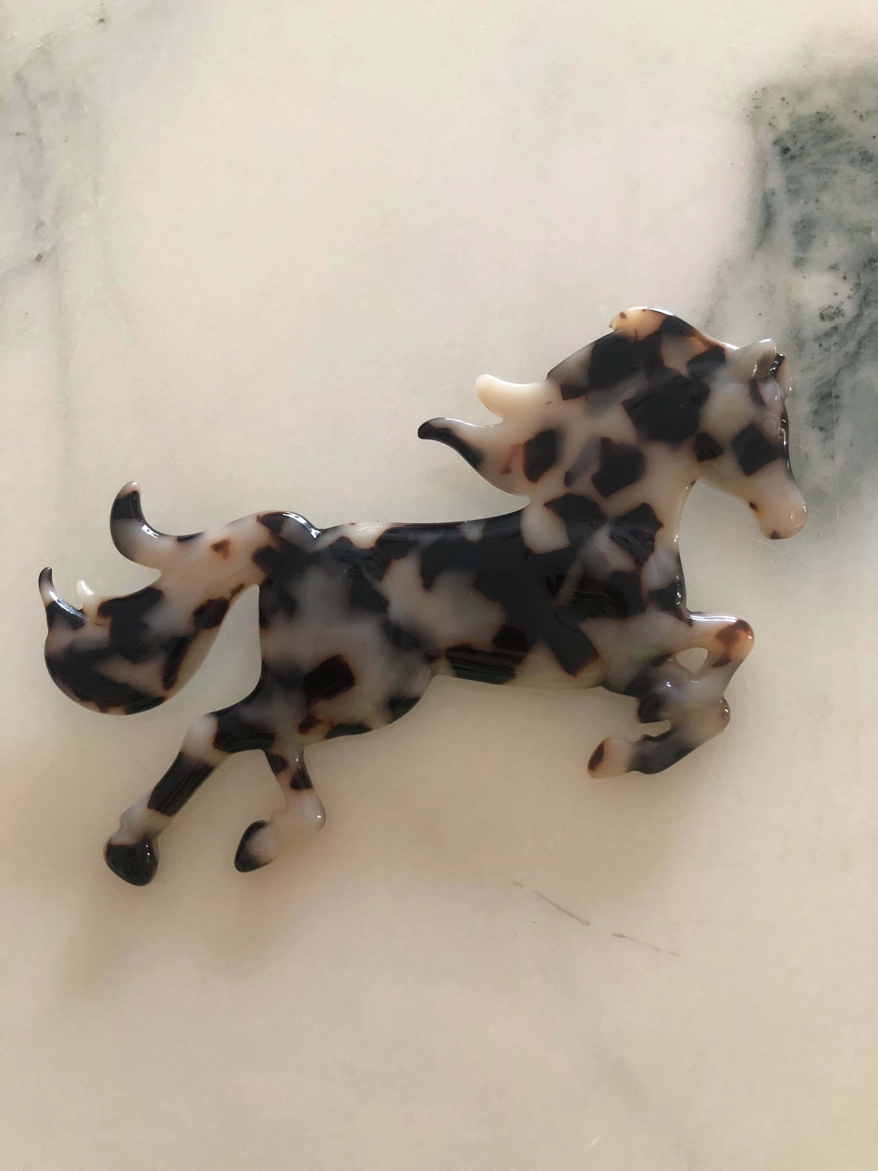 Broochella Men's Animalia  brown and black tortoiseshell figural horse brooch. It is displayed on a white marble surface. 