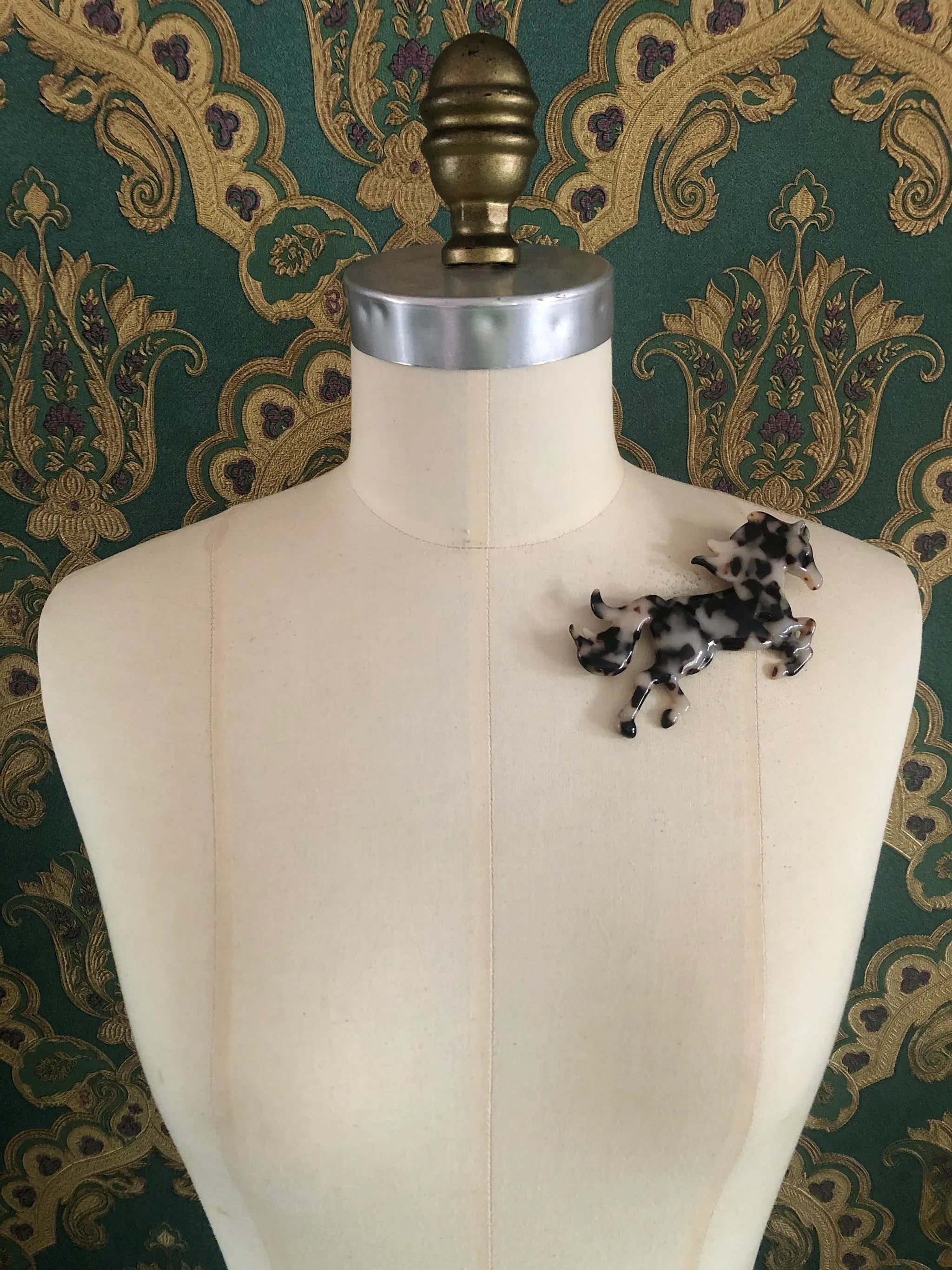 Broochella Animalia Collection beige, black and brown tortoiseshell horse brooch. It is made from premium, Italian cellulose acetate. The brooch is pinned to a mannequin. The background is green and gold damask wallpaper. 