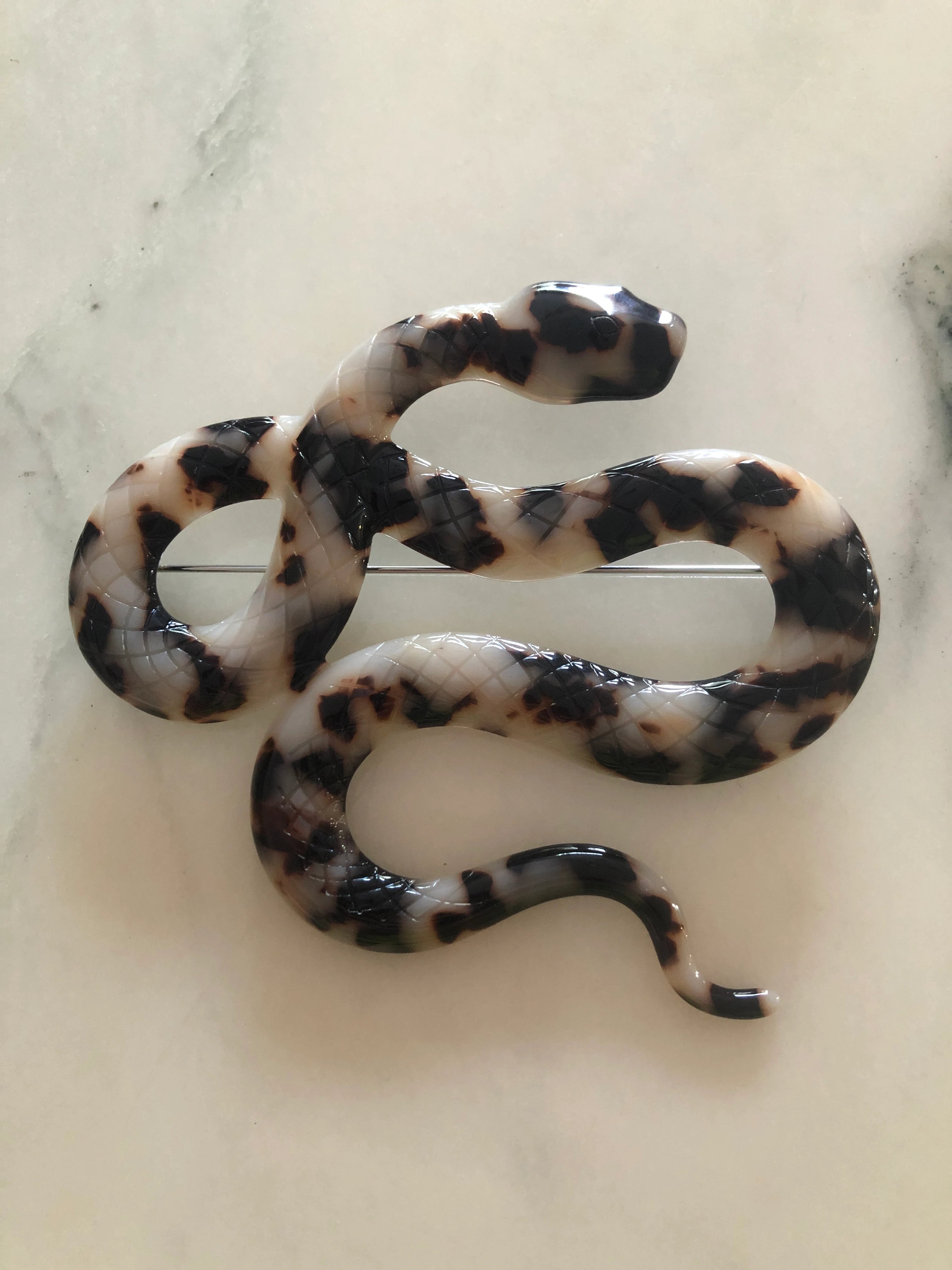 A Broochella Animalia beige, black and brown tortoiseshell snake-shaped brooch made from premium Italian cellulose acetate. It  displayed on a white marble table