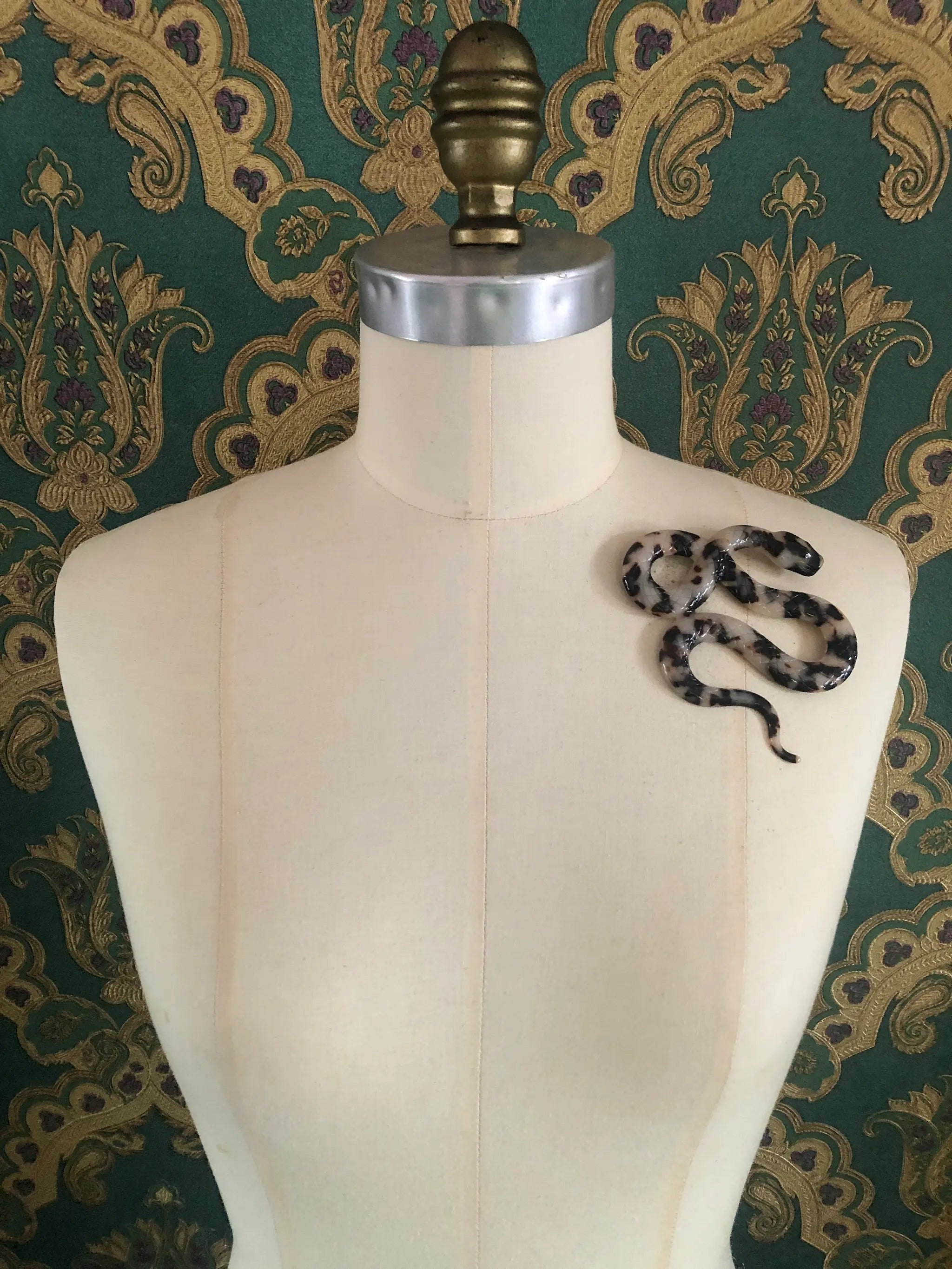A Broochella Animalia brown tortoiseshell snake-shaped brooch made from premium Italian cellulose acetate. It  displayed on a cream fabric dress form against a green and gold patterned wallpaper background.