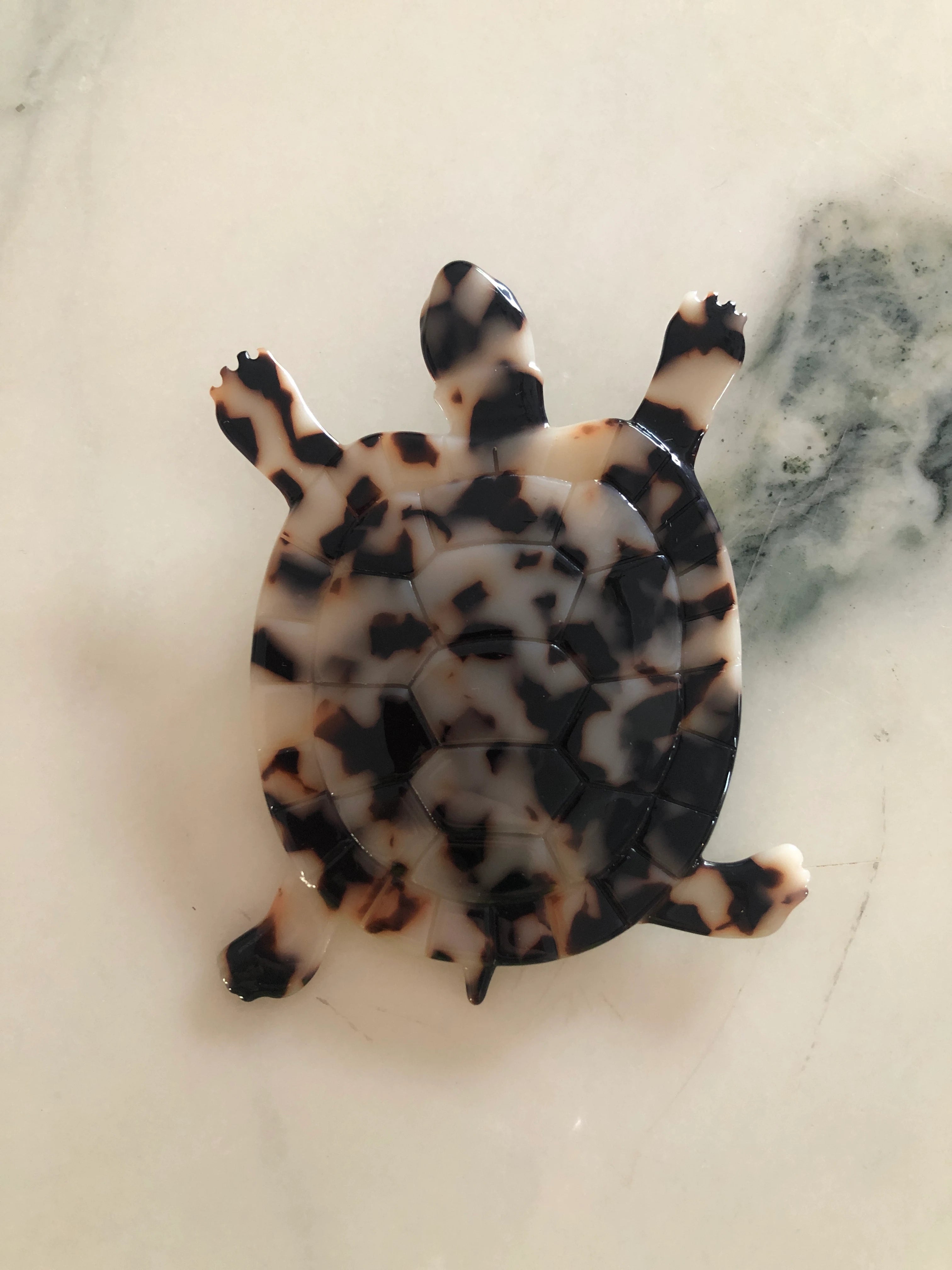 A Broochella Animalia Collection tortoise brooch in black, beige and brown tortoiseshell Italian cellulose acetate with detailed shell plate texture measuring 10cm x 8cm. The background is white marble. 