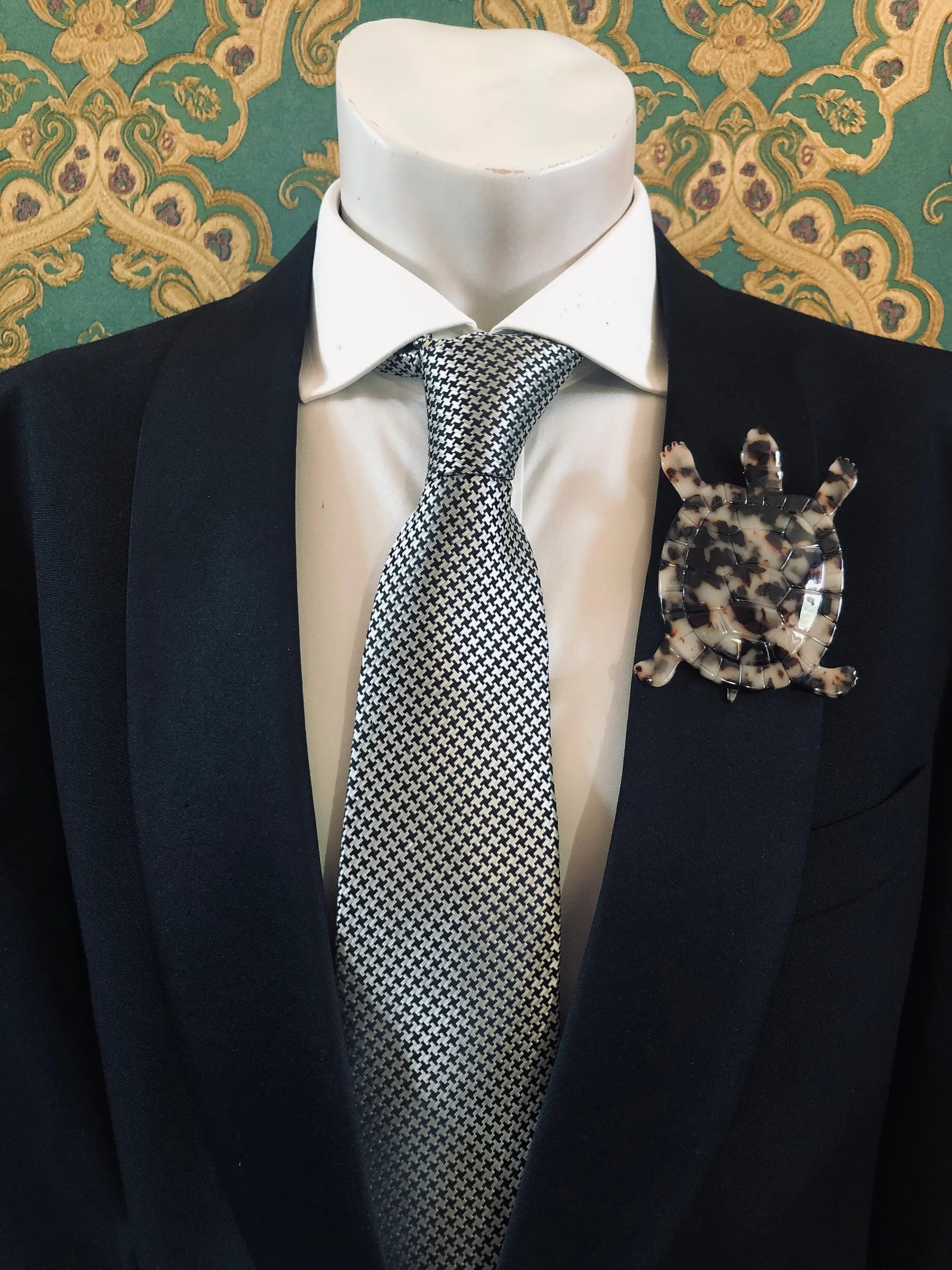 Broochella Men's beige Animalia tortoiseshell tortoise shaped brooch. It is made from Premium Italian cellulose tortoiseshell acetate. It is presented on a men's mannequin that is dressed in a black suit. The background is green and gold damask wallpaper. 