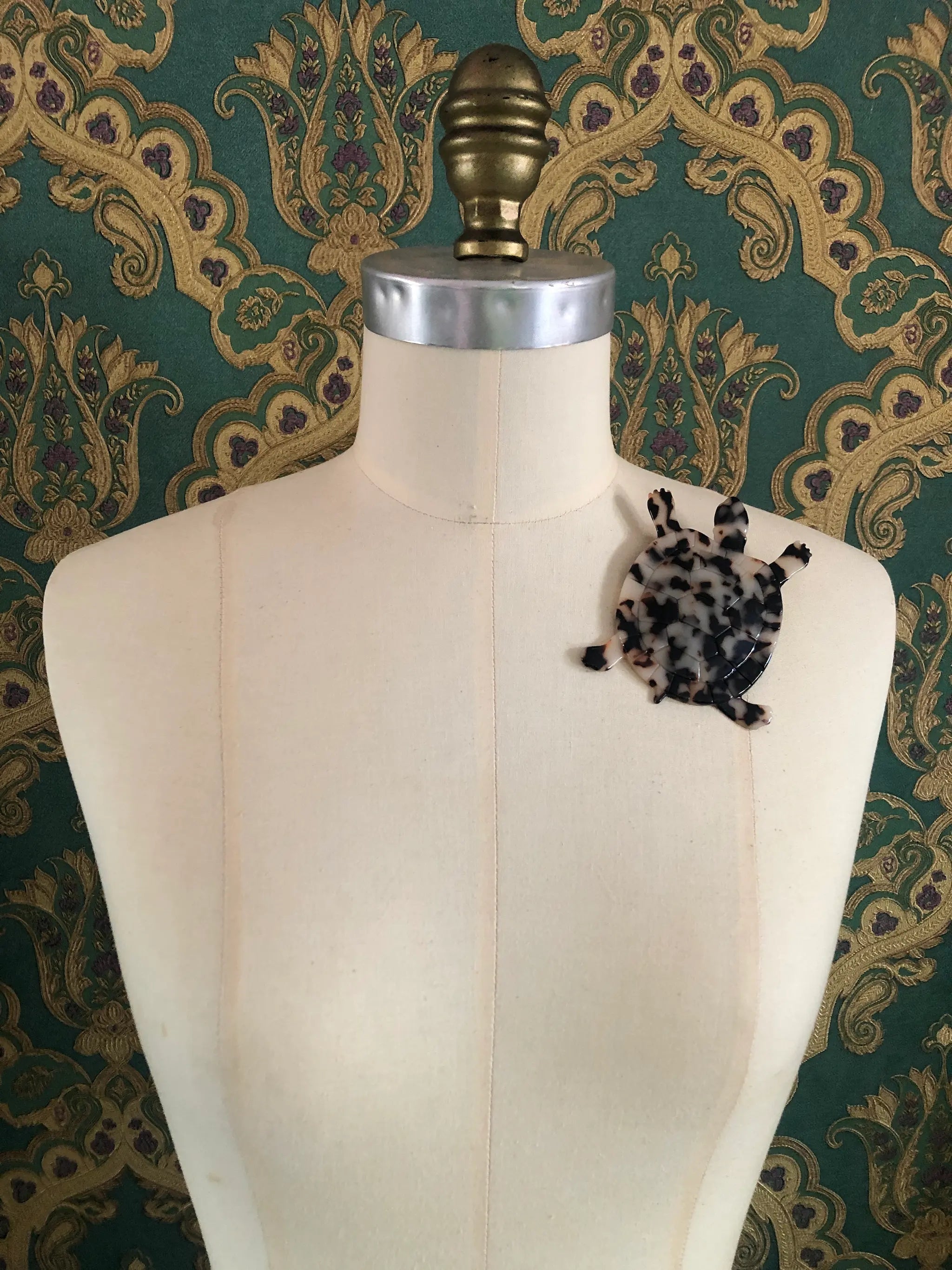 A Broochella Animalia Collection tortoise brooch in black, beige and brown tortoiseshell Italian cellulose acetate with detailed shell plate texture measuring 10cm x 8cm. It is pinned to a mannequin. The background is green and gold damask wallpaper. 