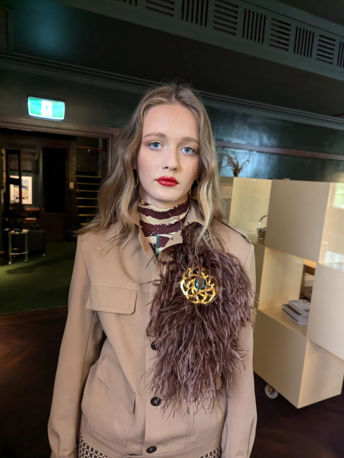 A woman with blonde hair and blue eyes wearing a beige coat over a brown Broochella bejewelled feathered bow brooch with a gold brooch embellishment standing in an indoor setting.