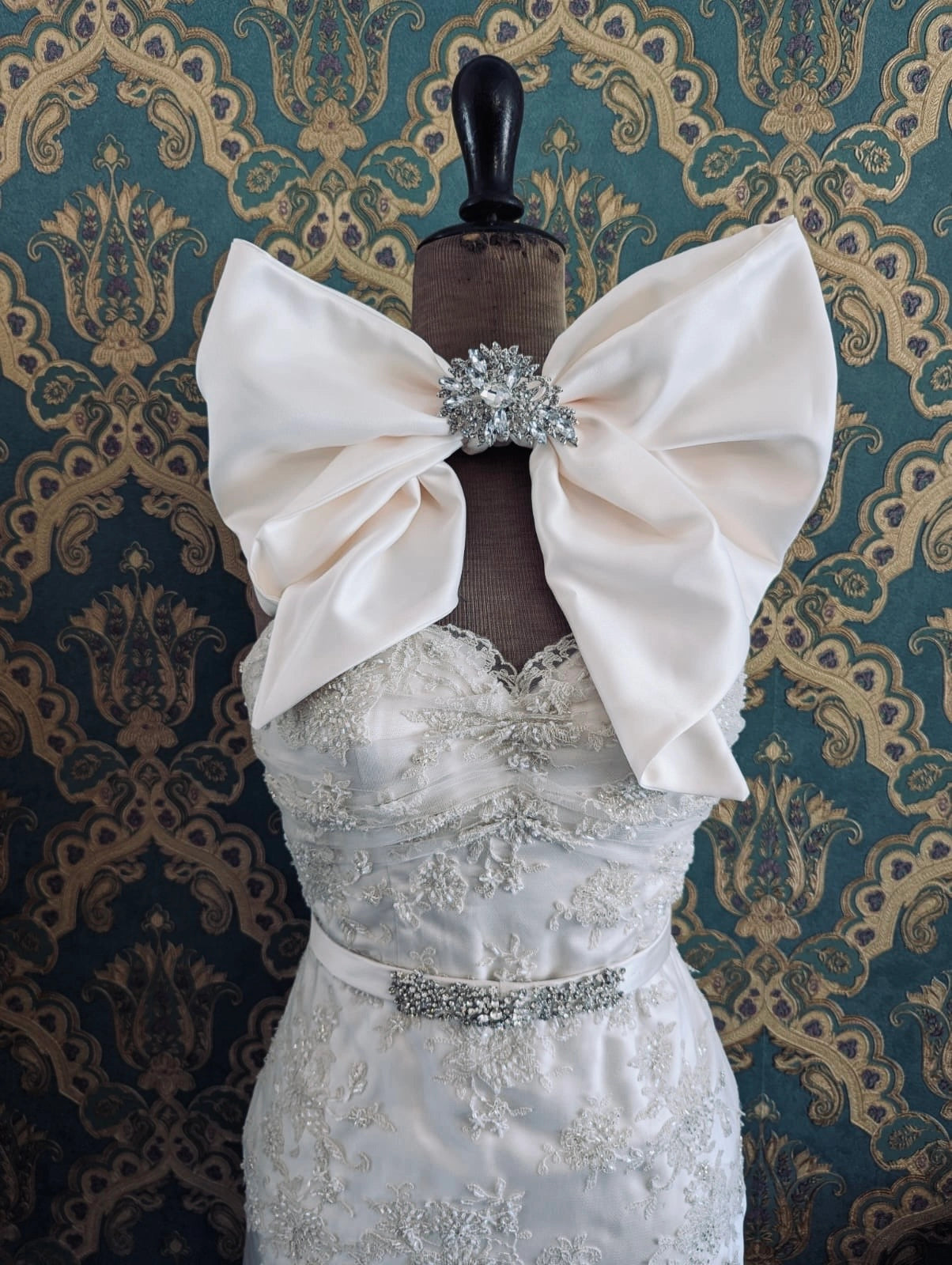 Broochella bejewelled large, bridal, handmade satin bow, with crystal brooch embellishment. Pinned on a mannequin. The mannequin is dressed in a white, beaded, strapless wedding dress. The background is green and gold damask wallpaper.