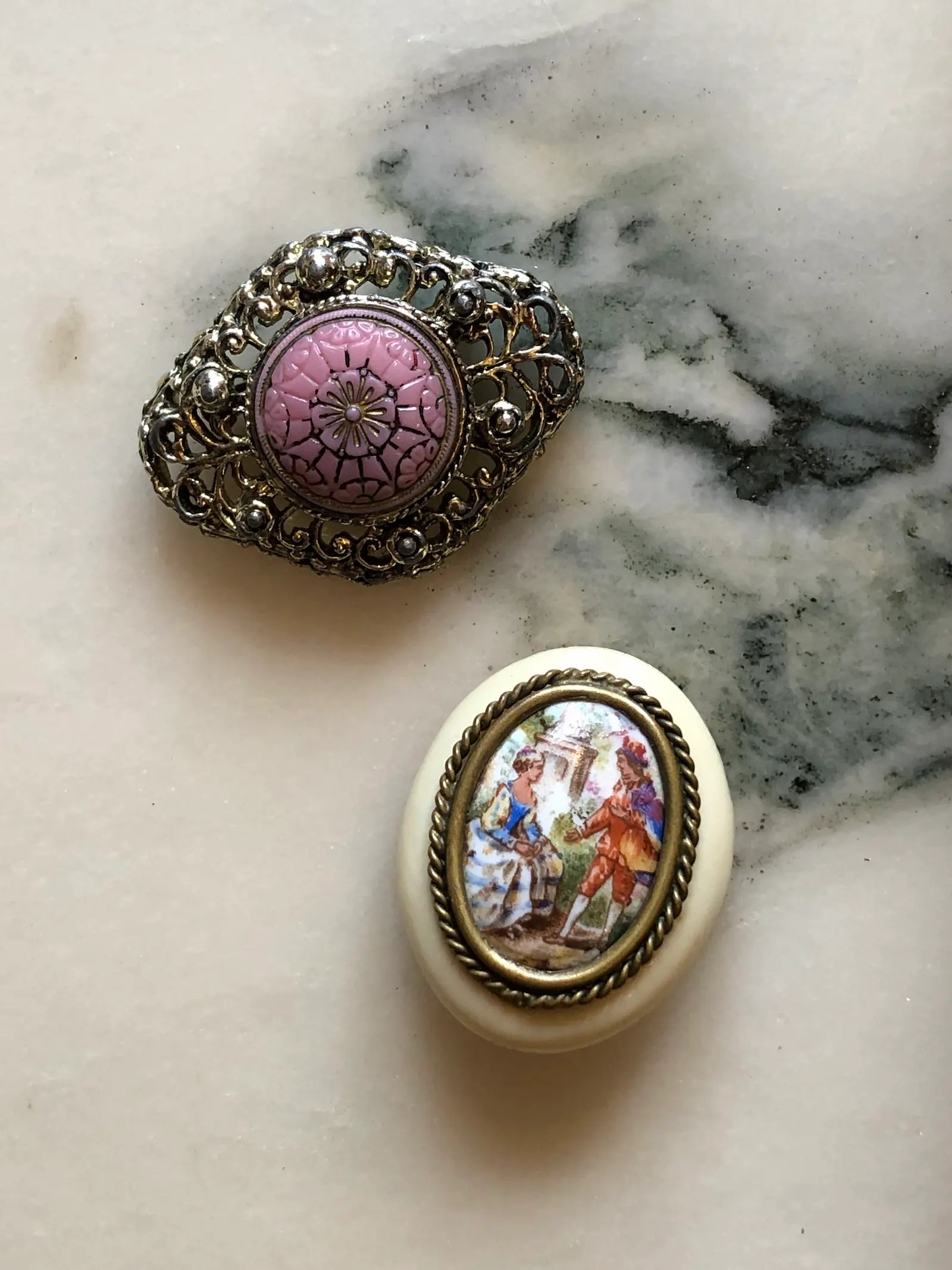 Broochella Belle Époque vintage brooch duo with a pink floral filigree brooch and a French Limoges porcelain courting-scene brooch. The background is white 