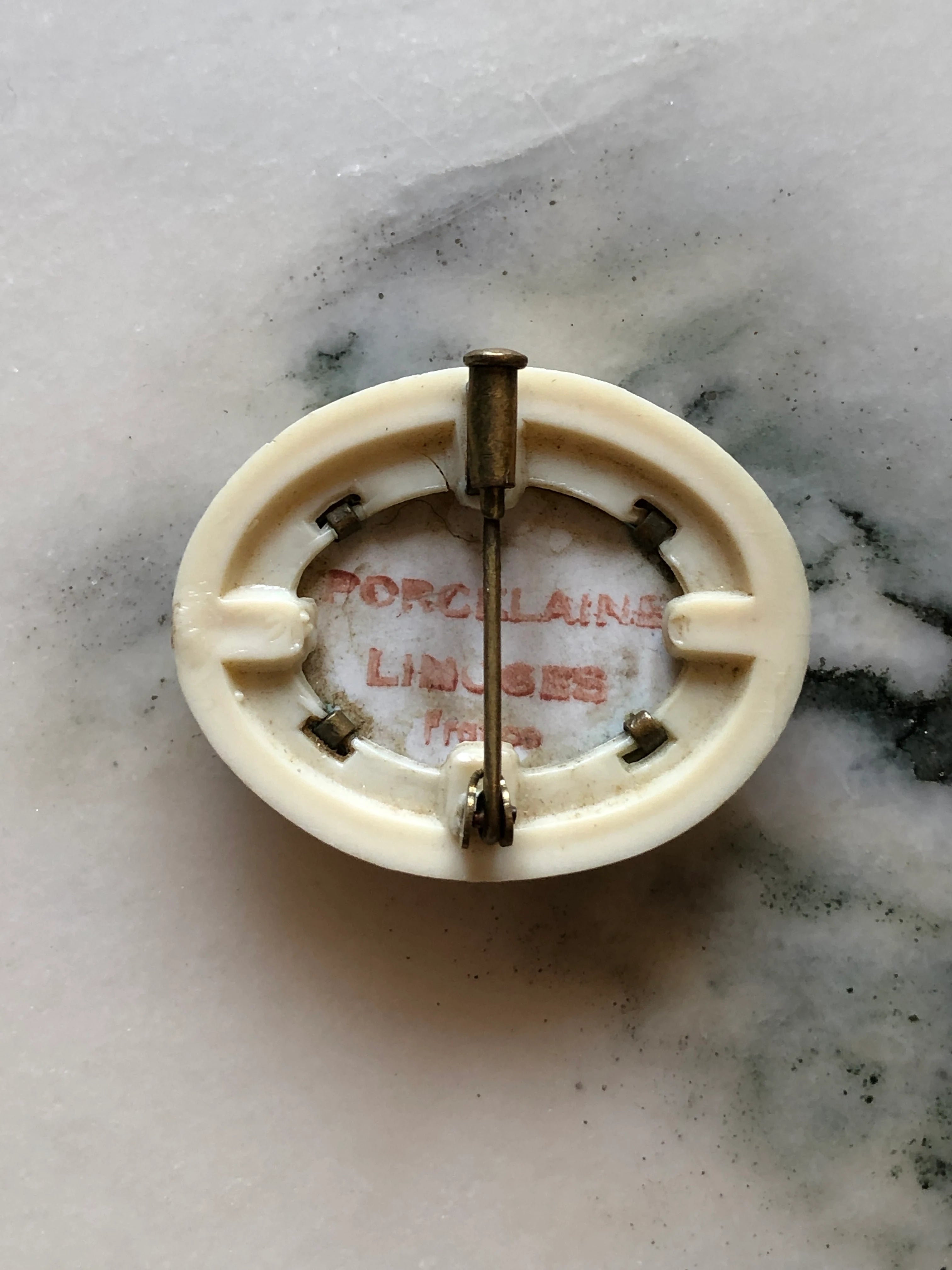 A Broochella Belle Epoque oval-shaped cream-colored porcelain pin back with a metal safety pin attached, resting on a white and gray marble surface. The inside of the porcelain is stamped with 'PORCELAIN LIMOGES Paris'.
