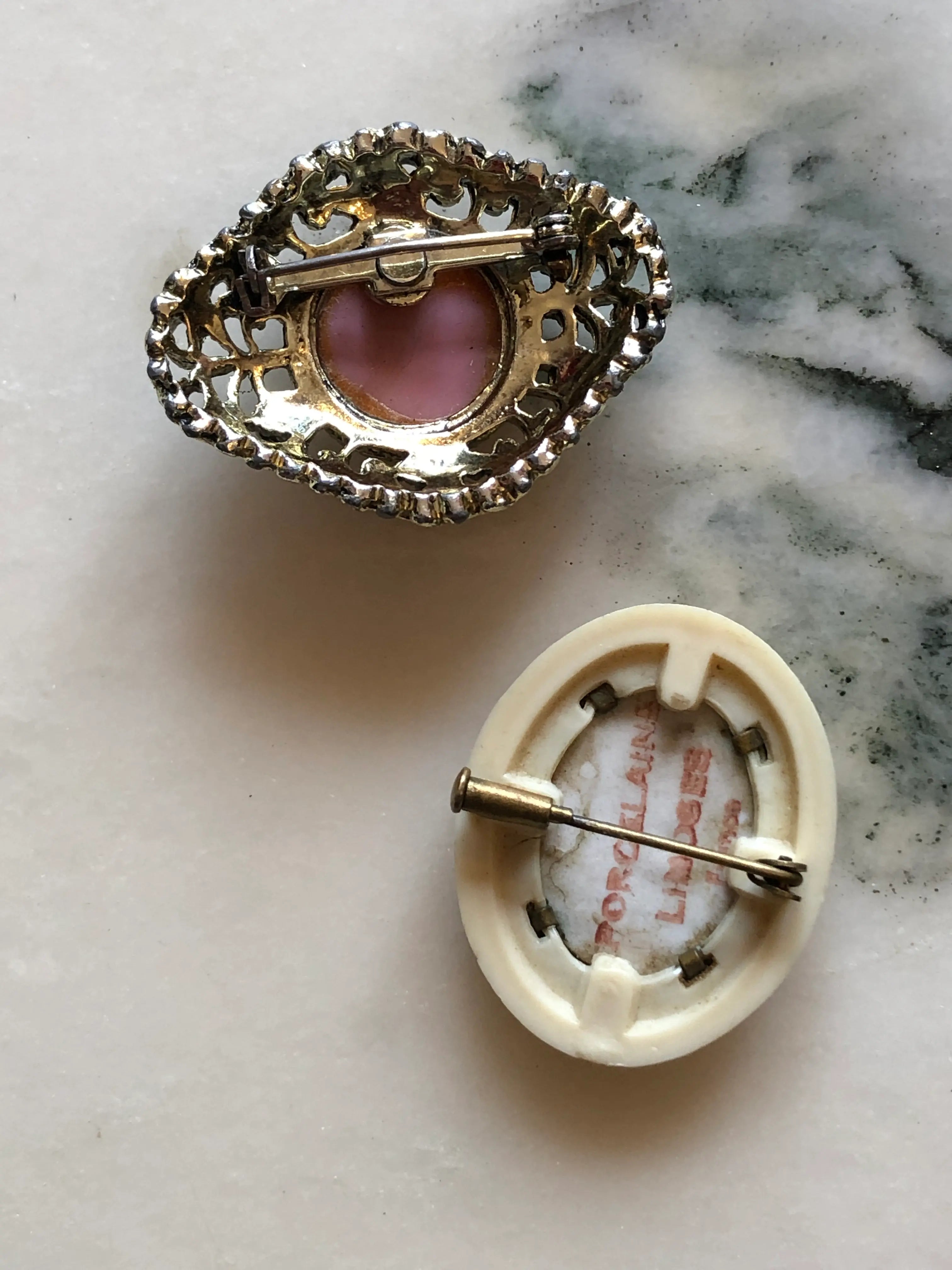 The back view of Broochella Belle Époque vintage brooch duo with a pink floral filigree brooch and a French Limoges porcelain courting-scene brooch. The background is white marble.