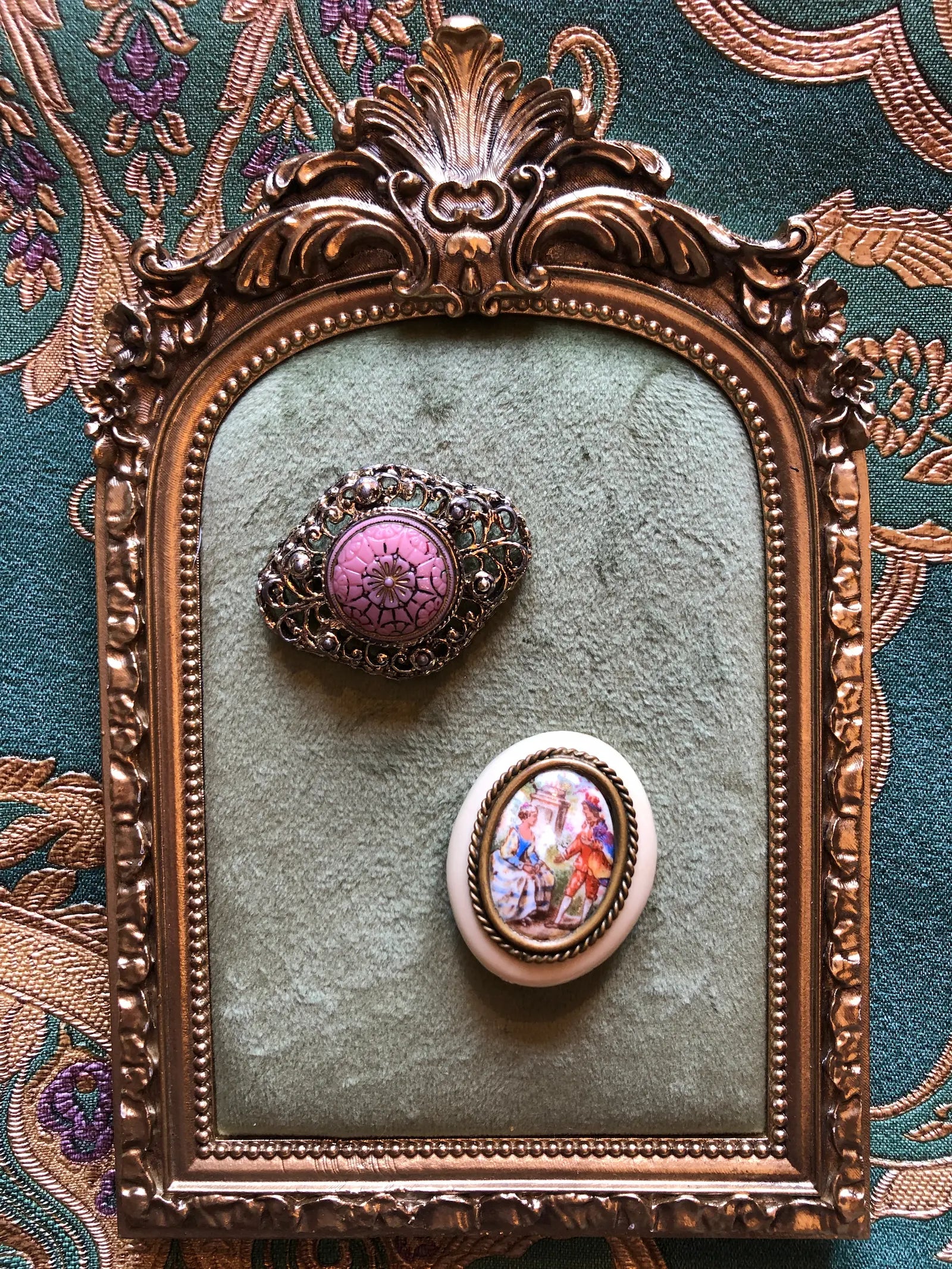 Broochella Belle Époque vintage brooch duo with a pink floral filigree brooch and a French Limoges porcelain courting-scene brooch. They are displayed on a green velvet insert inside an ornate gold frame, set against a patterned teal fabric background.