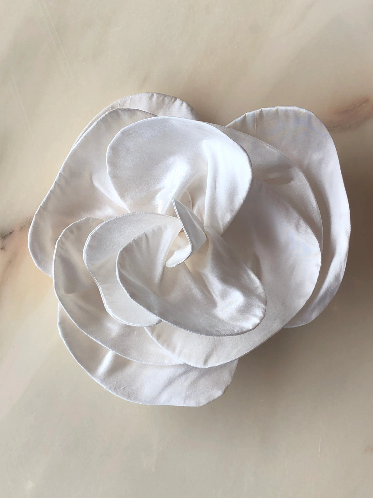 Broochella Bellucci Bridal Brooch in white, featuring an oversized sculptural silk flower with softly curved layered petals, photographed on a pale cream marble background.