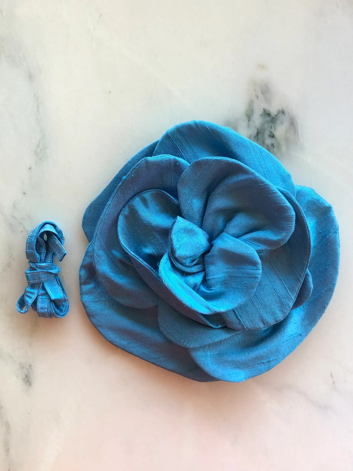 Alt text: Broochella Bellucci Large Silk Flower Brooch & Choker in vibrant blue, with sculptural layered silk petals and matching ribbon, photographed on a white marble background with soft grey veining