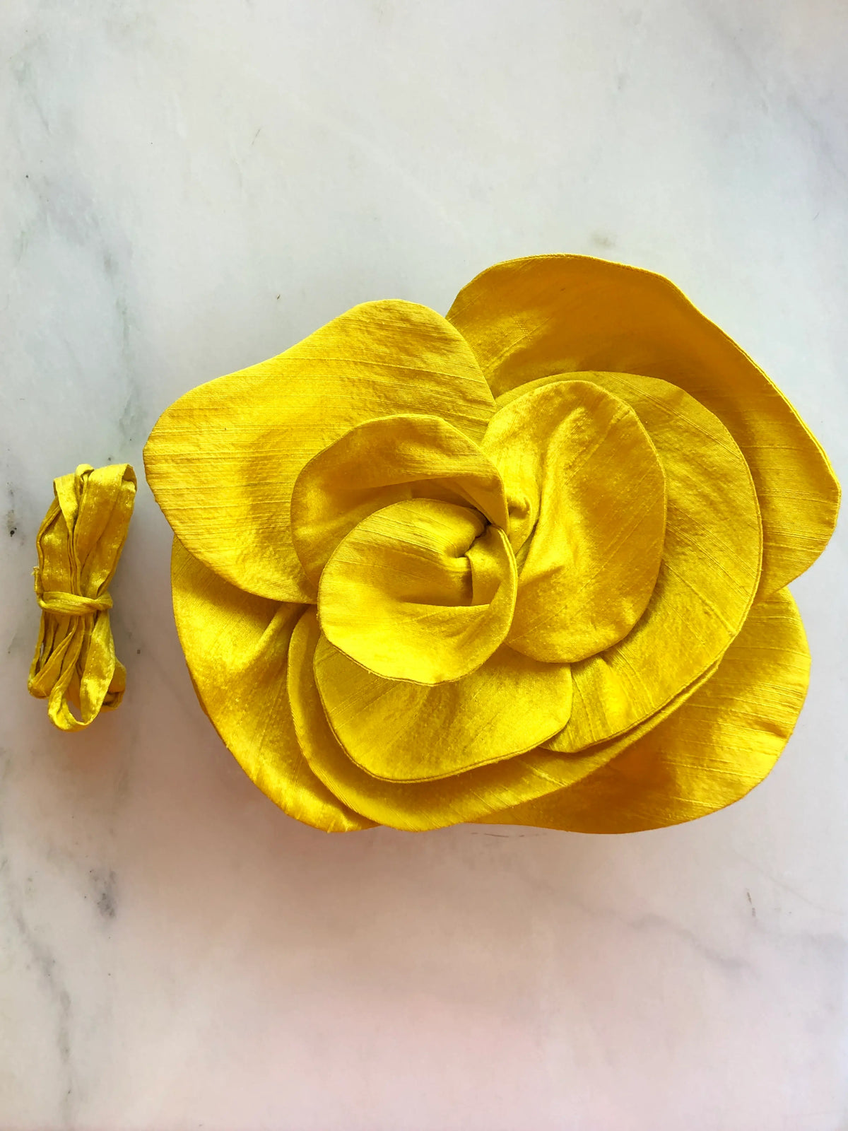 Broochella Bellucci Large Silk Flower Brooch & Choker in vibrant yellow, with sculptural layered silk petals and matching ribbon, photographed on a white marble background with subtle grey veining.