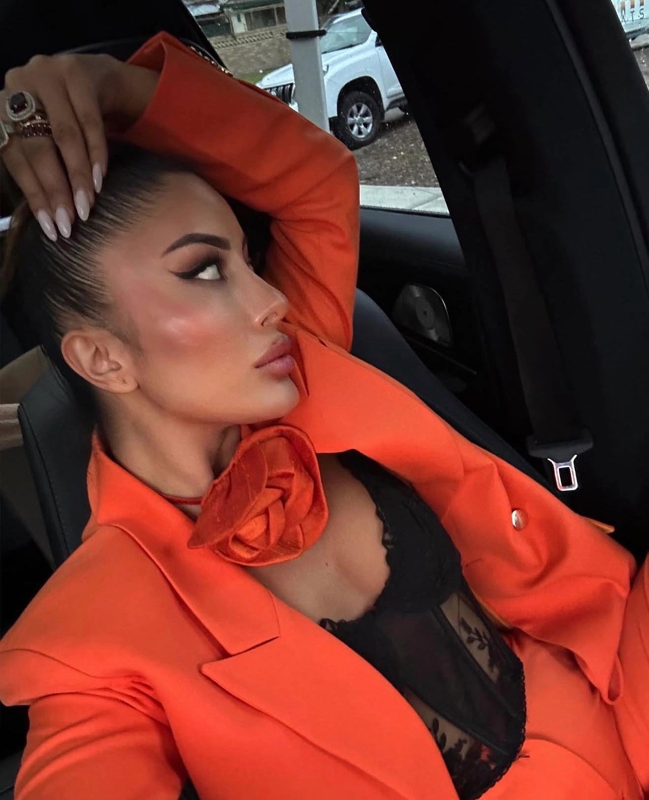 A woman posing inside a car wearing a bright orange satin blazer with a large matching Broochella Bellucci  orange flower brooch choker  accessory at the collar, layered over a black lace top.
