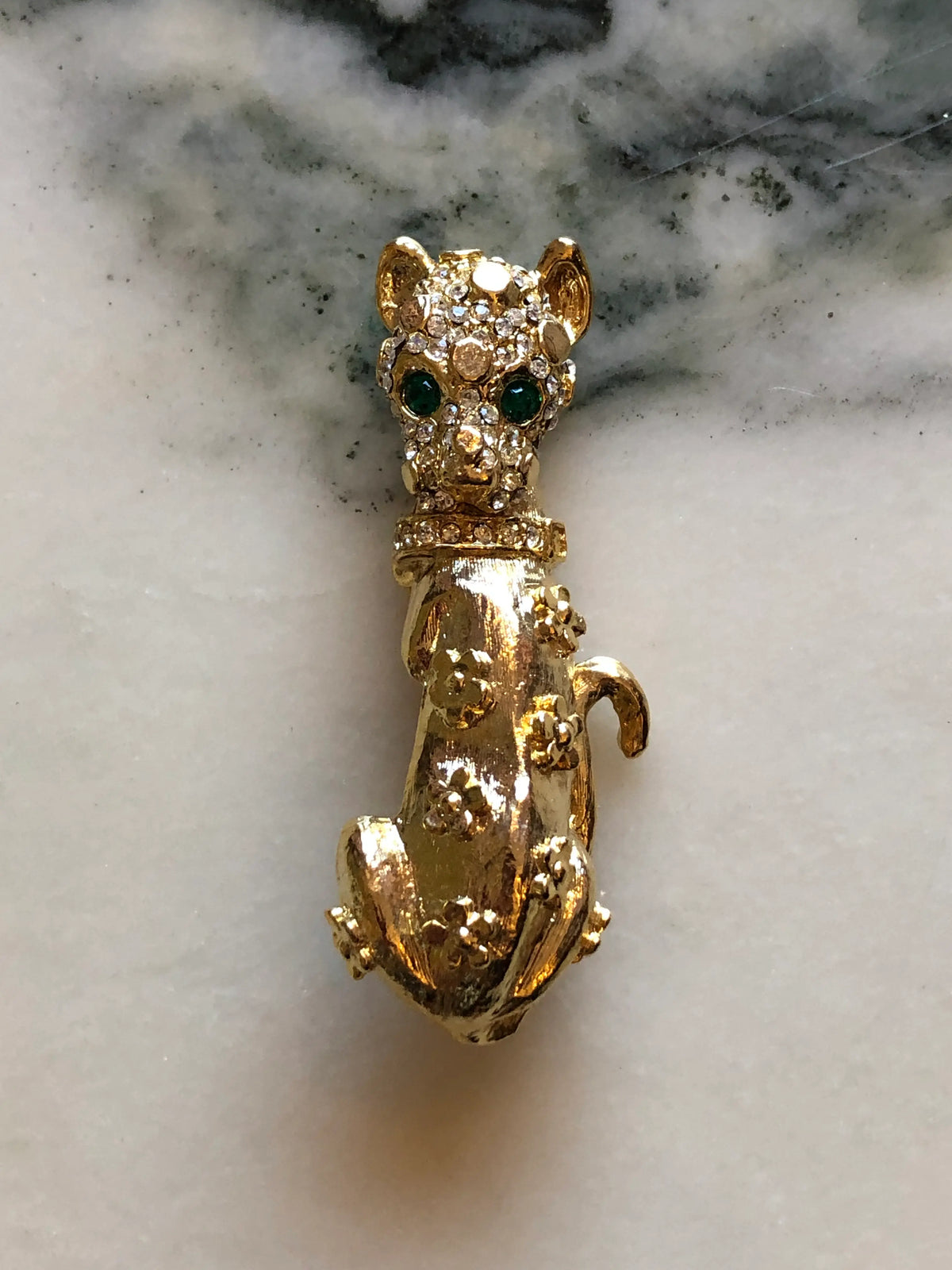 Broochella Bianca Vintage Gold Cat Brooch featuring an elongated gold-tone cat with emerald-green eyes, clear crystal detailing and floral accents on a cream and grey marble background.