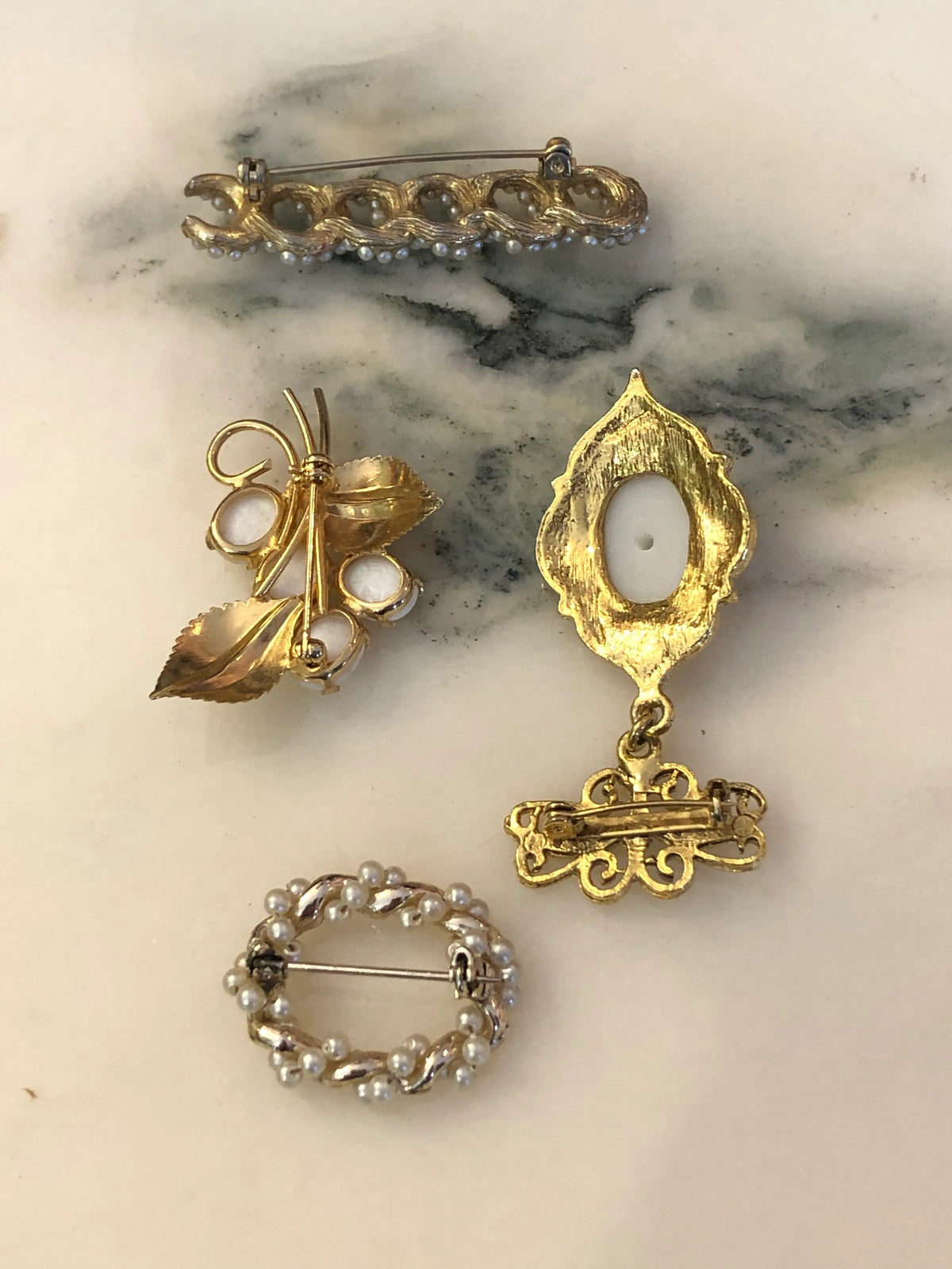 The back view of Broochella The Bianca Vintage Brooch Quartet featuring four ornate vintage pearl, ivory and gold-tone brooches with botanical, scroll and filigree details on a cream and grey marble background.