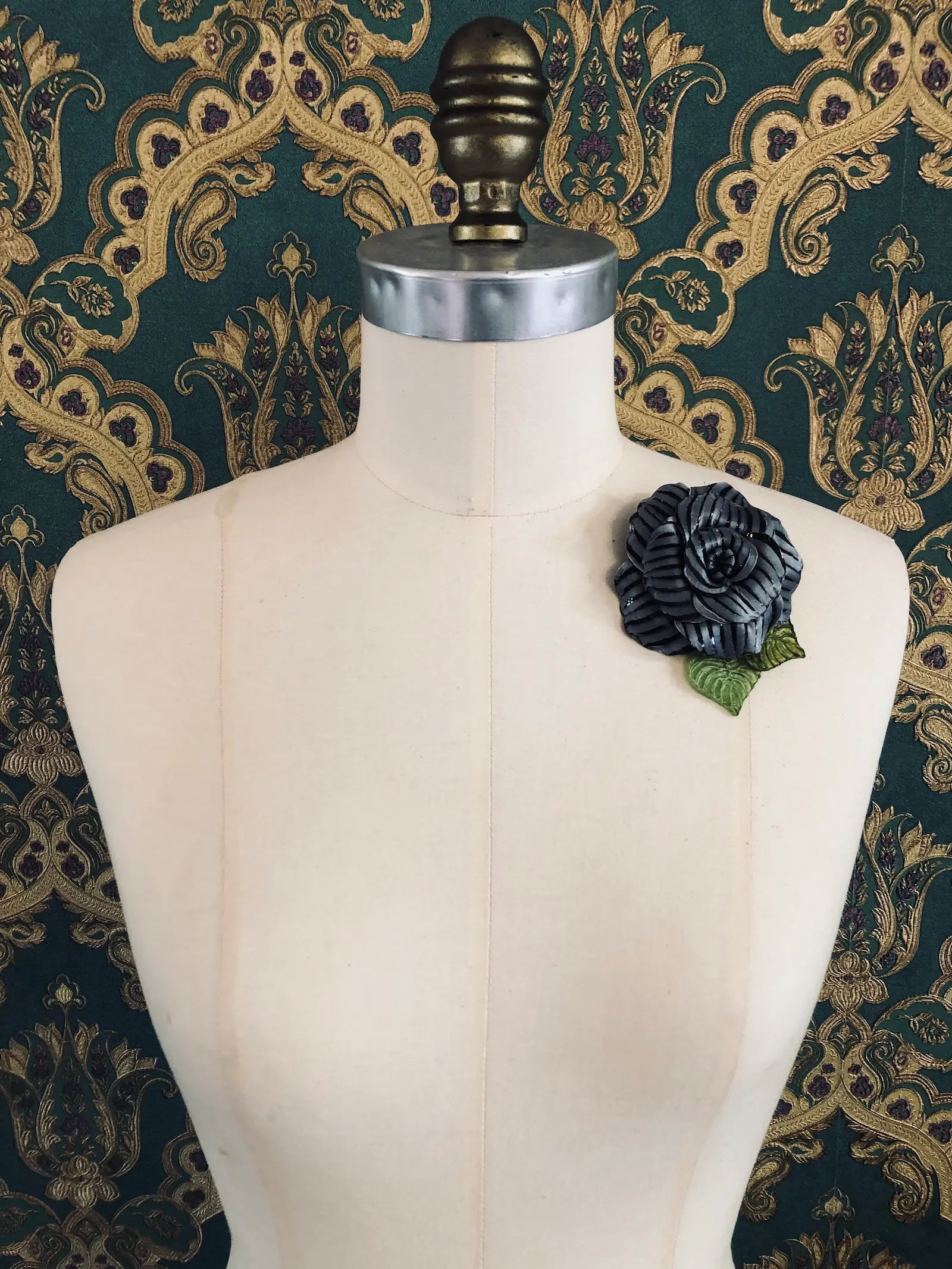 A Broochella x Fabrice Paris black moulded plastic rose brooch with green leaves, pinned to the chest of a cream-coloured dress form against a dark green and gold patterned damask backdrop.