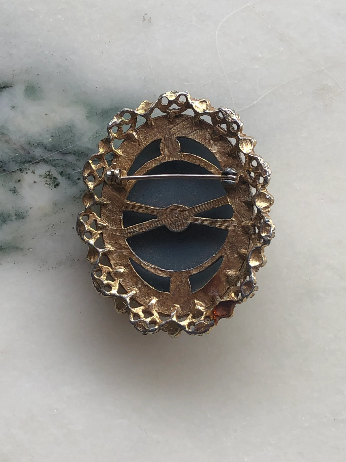 The back view of Broochella bleu vintage cameo brooch. It features a gold decorative frame and a blue and white cameo with the profile of a woman with long hair holding a flower. The background is white marble
