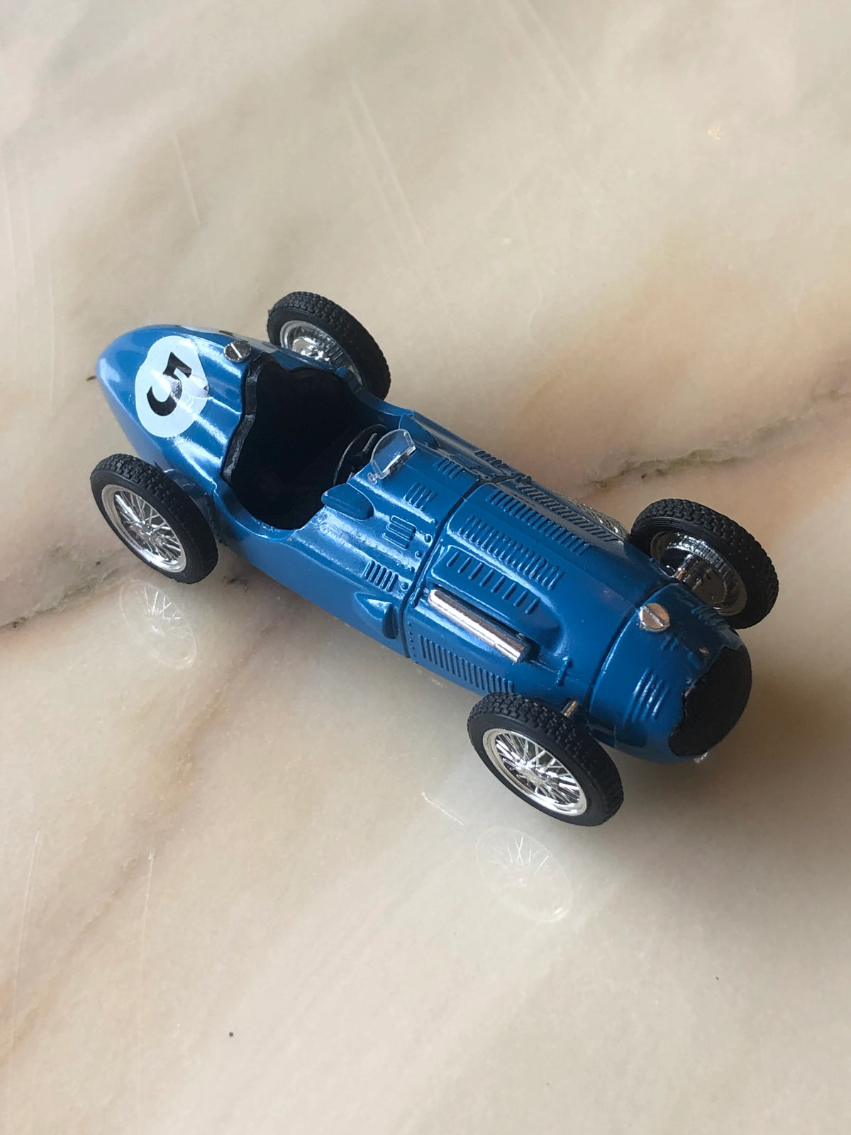 Broochella Blu Veloce No.5 Vintage Grand Prix Racing Car Brooch in glossy blue, featuring a black open cockpit, silver wire-spoke wheels, detailed vented bonnet and white number 5 racing decal, photographed from an elevated three-quarter angle.