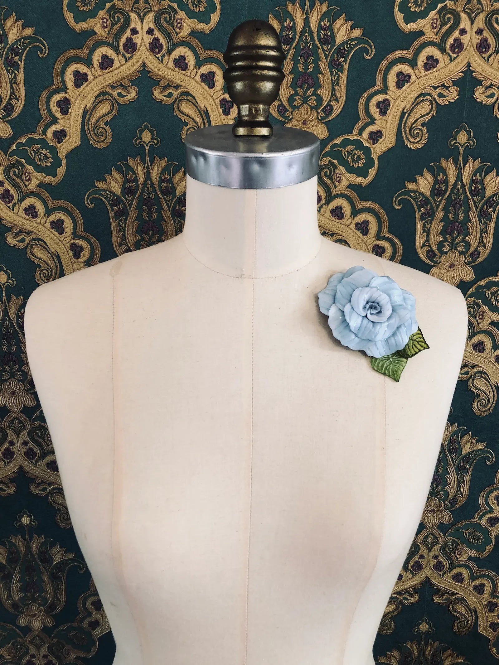 A Broochella x Fabrice Paris light blue moulded plastic rose brooch with green leaves, pinned to the chest of a cream-colored dress form against a dark green and gold patterned damask backdrop.