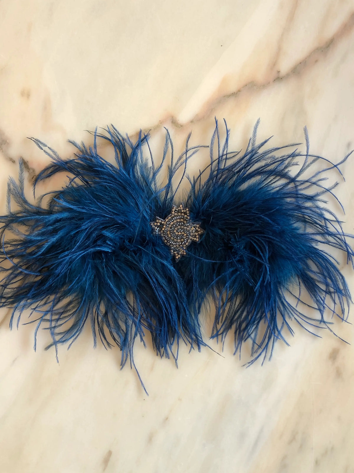 Broochella handmade Blue Feather Bow Brooch featuring dramatic ostrich feathers and an ornate jewelled centre, an oversized designer statement brooch photographed on a white marble background.