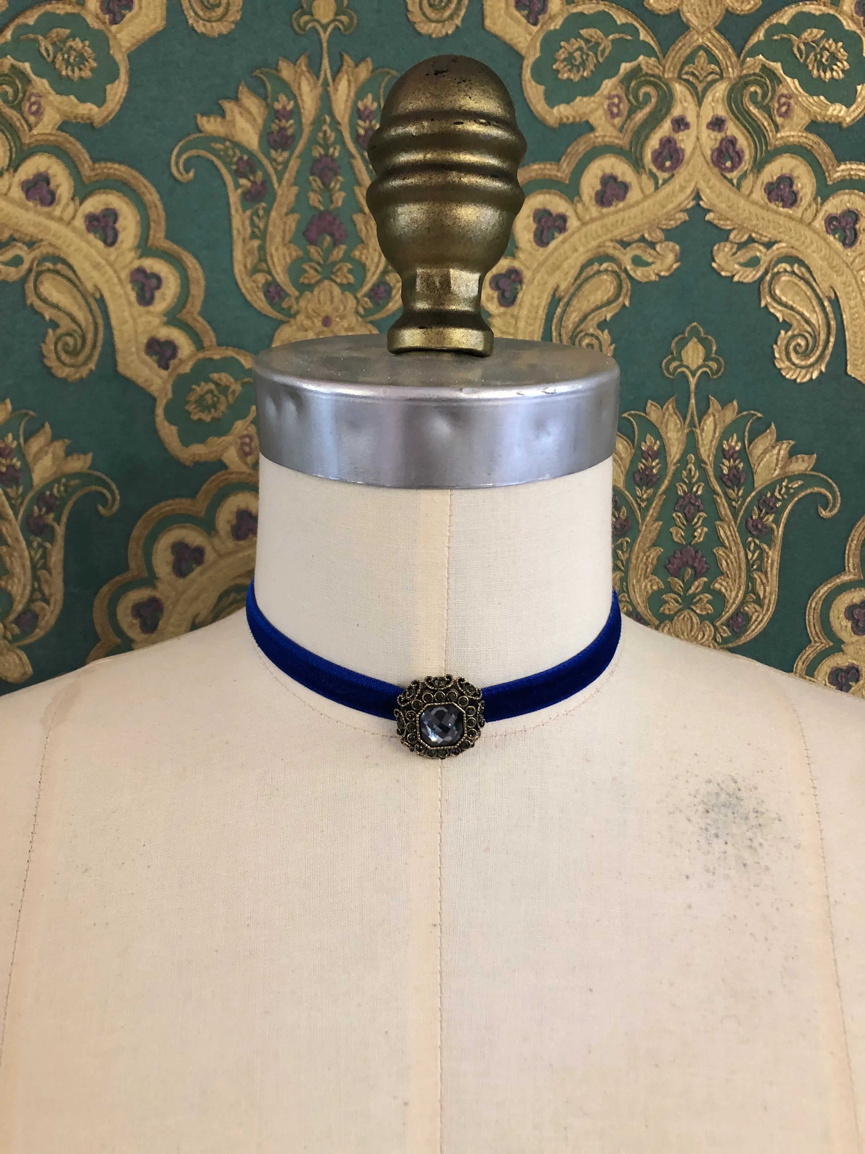 A Broochella Lady Whistledown blue velvet choker necklace with a decorative antique bronze filigree centrepiece, displayed on a cream-coloured dress form against a patterned teal and gold damask wallpaper background.