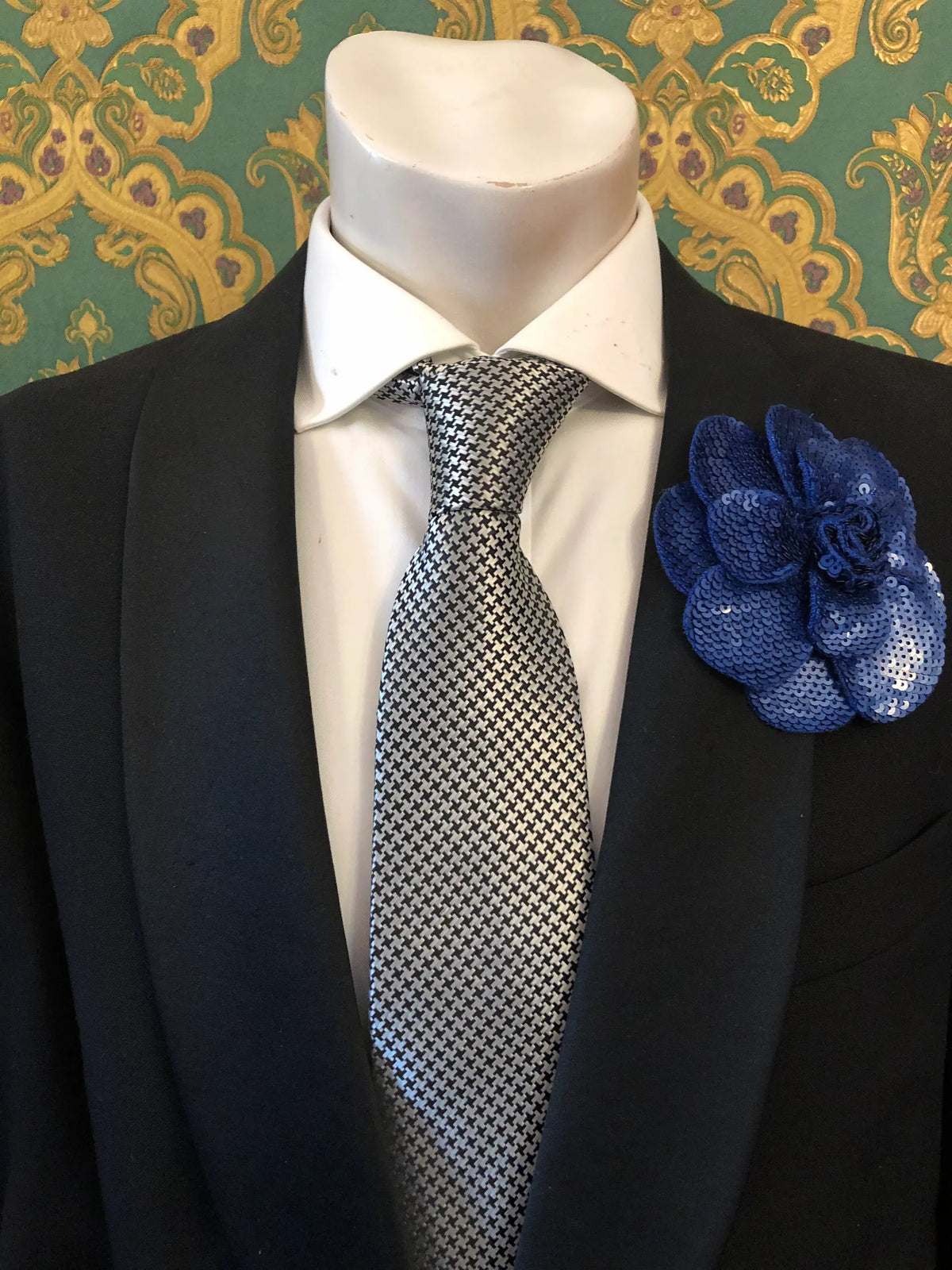 A men's formal accessory set displayed on a white mannequin torso, featuring a black suit jacket, a white dress shirt, a silver and black checkered tie, and an orange Men's Broochella Scintillante sequinned flower brooch lapel pin against a green and gold patterned wallpaper background.