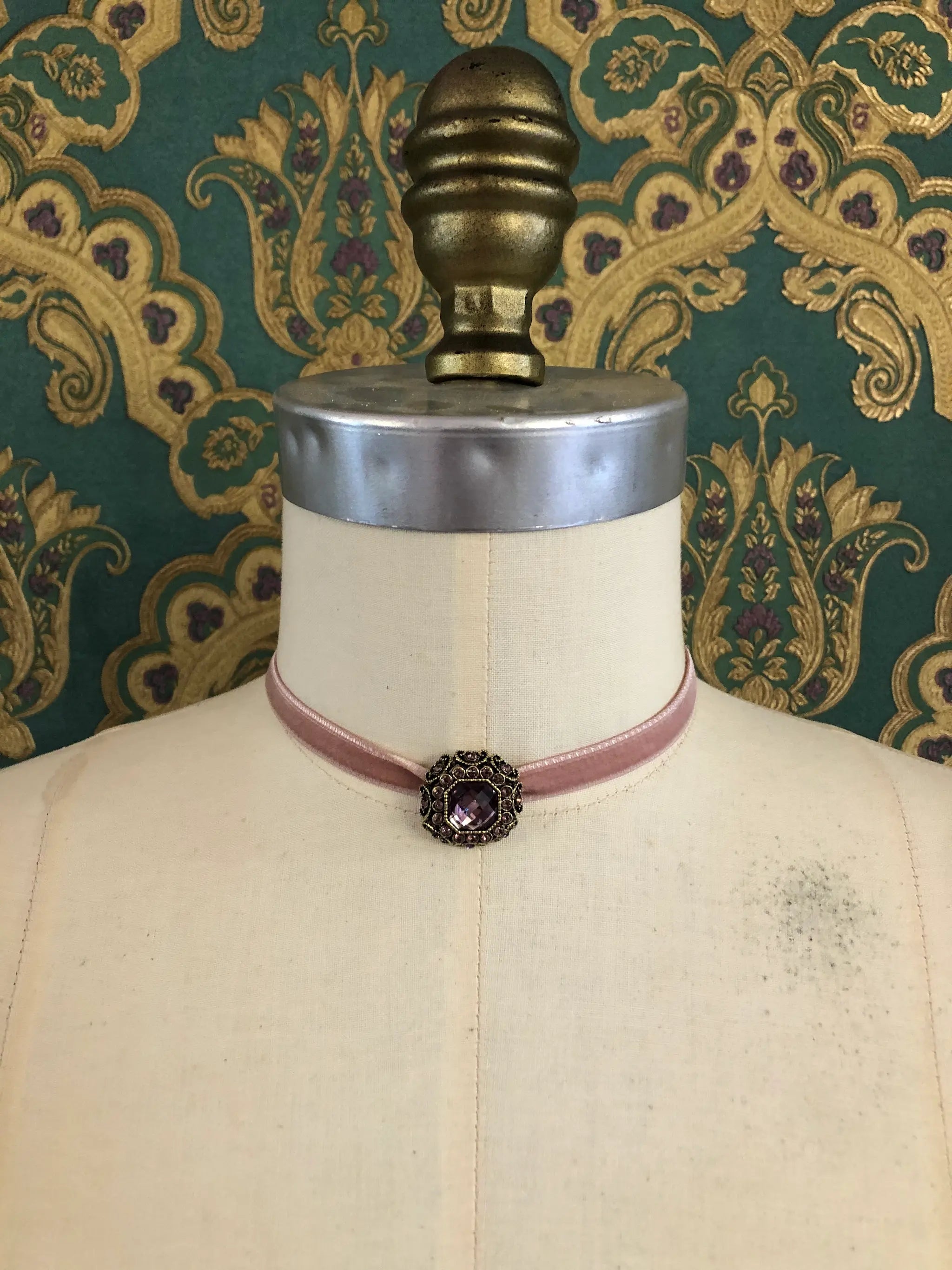 A Broochella Lady Whistledown blush pink velvet choker necklace with a decorative antique bronze filigree centrepiece, displayed on a cream-coloured dress form against a patterned teal and gold damask wallpaper background.
