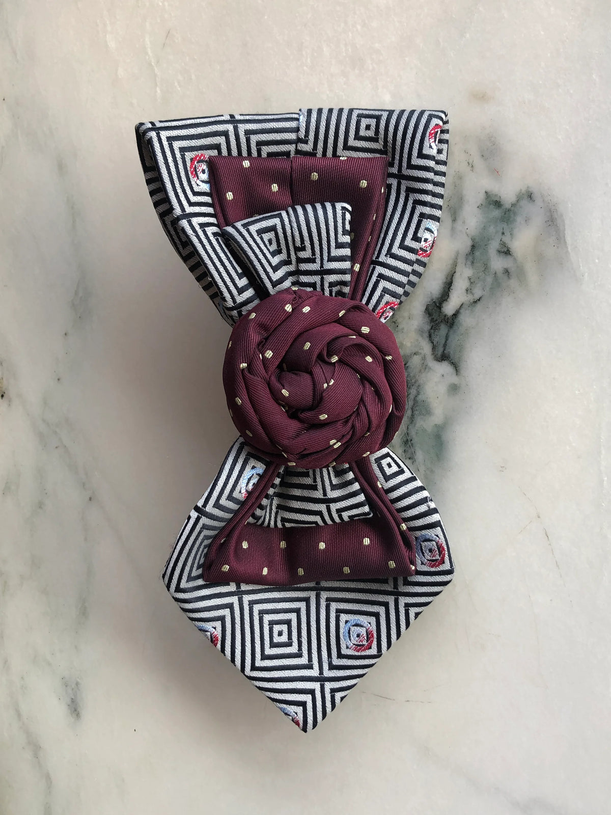 Broochella Bolivar Cravatte Tie Brooch Pin No. 10, a one-of-a-kind statement brooch handmade from repurposed vintage ties in burgundy, black and white geometric patterns, finished with a sculptural burgundy rosette centre, photographed vertically on white marble with grey-green veining.