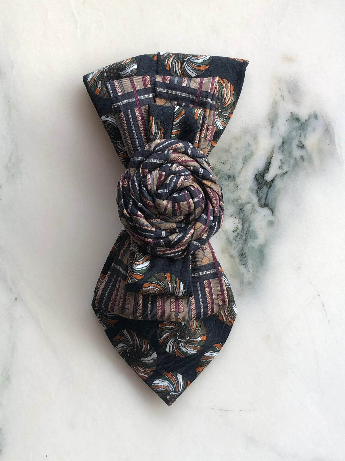 Broochella Bolivar Cravatte Tie Brooch Pin No. 2, a one-of-a-kind statement brooch handmade from repurposed vintage patterned ties in black, brown, burgundy and cream, finished with a sculptural knotted rosette centre and photographed on white marble with grey-green veining.