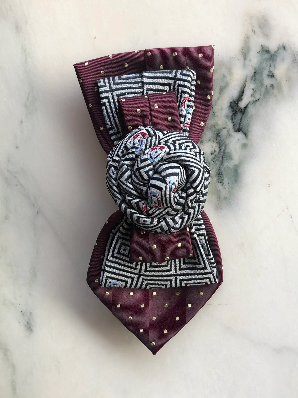 Broochella Bolivar Cravatte Tie Brooch Pin No. 9, a one-of-a-kind statement brooch handmade from repurposed vintage ties in burgundy, black and white geometric silk, with a sculptural knotted rosette centre, photographed on white marble with grey-green veining.