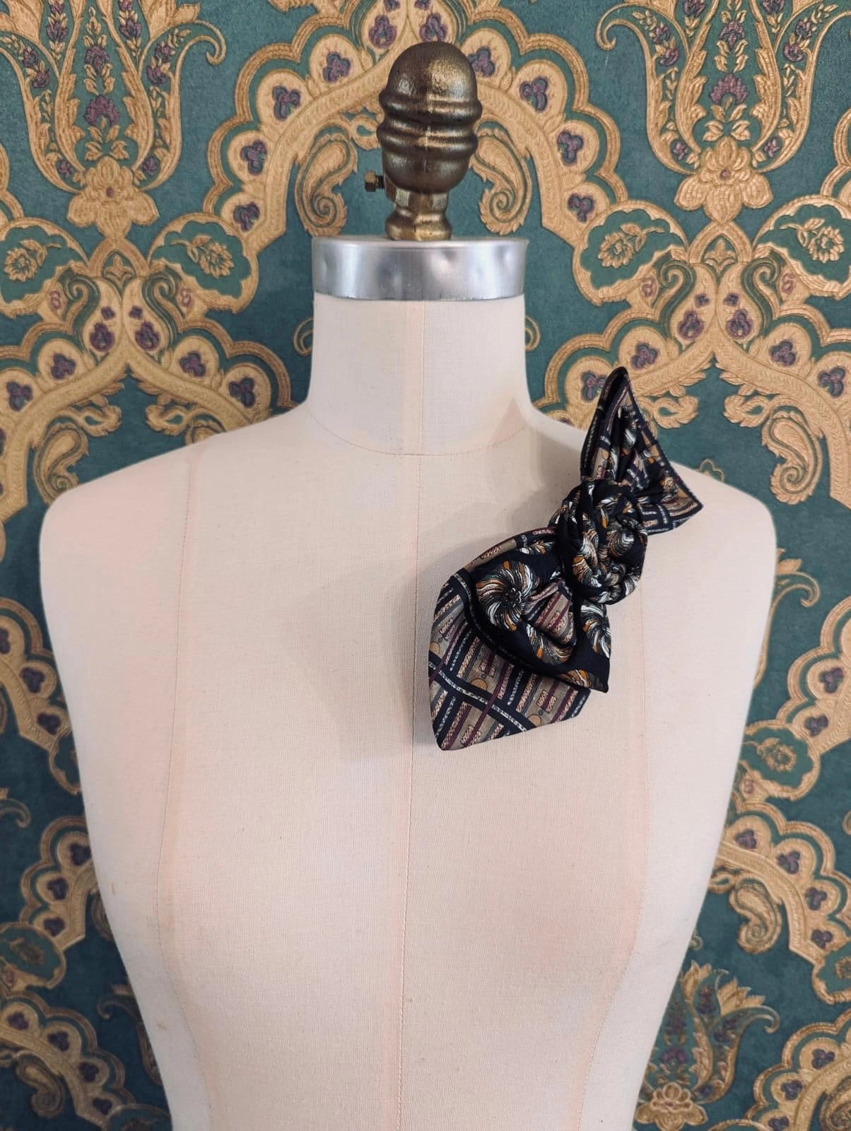 A Broochella Bolivar vintage upcycled necktie styled on a cream fabric dress form mannequin, featuring a black and brown geometric pattern with a central fabric rose embellishment. The background is green and gold damask wallpaper.