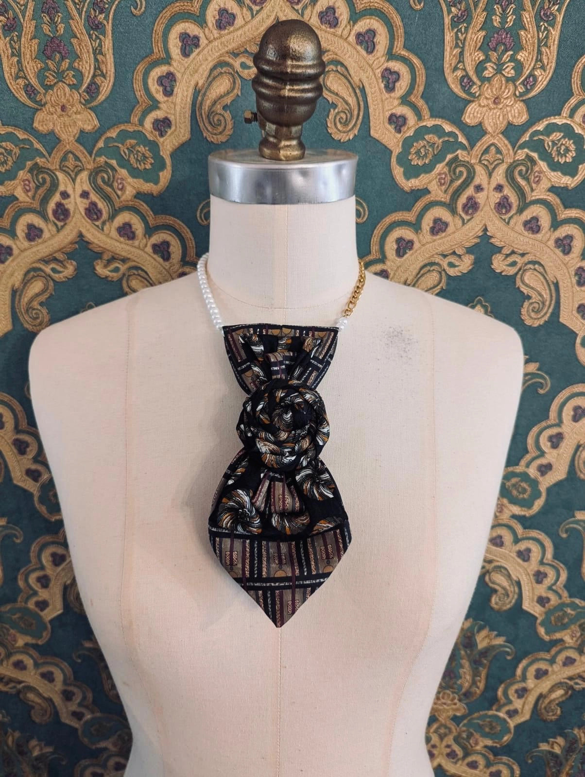 A Broochella Bolivar vintage upcycled necktie styled on a cream fabric dress form mannequin, featuring a black and brown geometric pattern with a central fabric rose embellishment. The background is green and gold damask wallpaper.