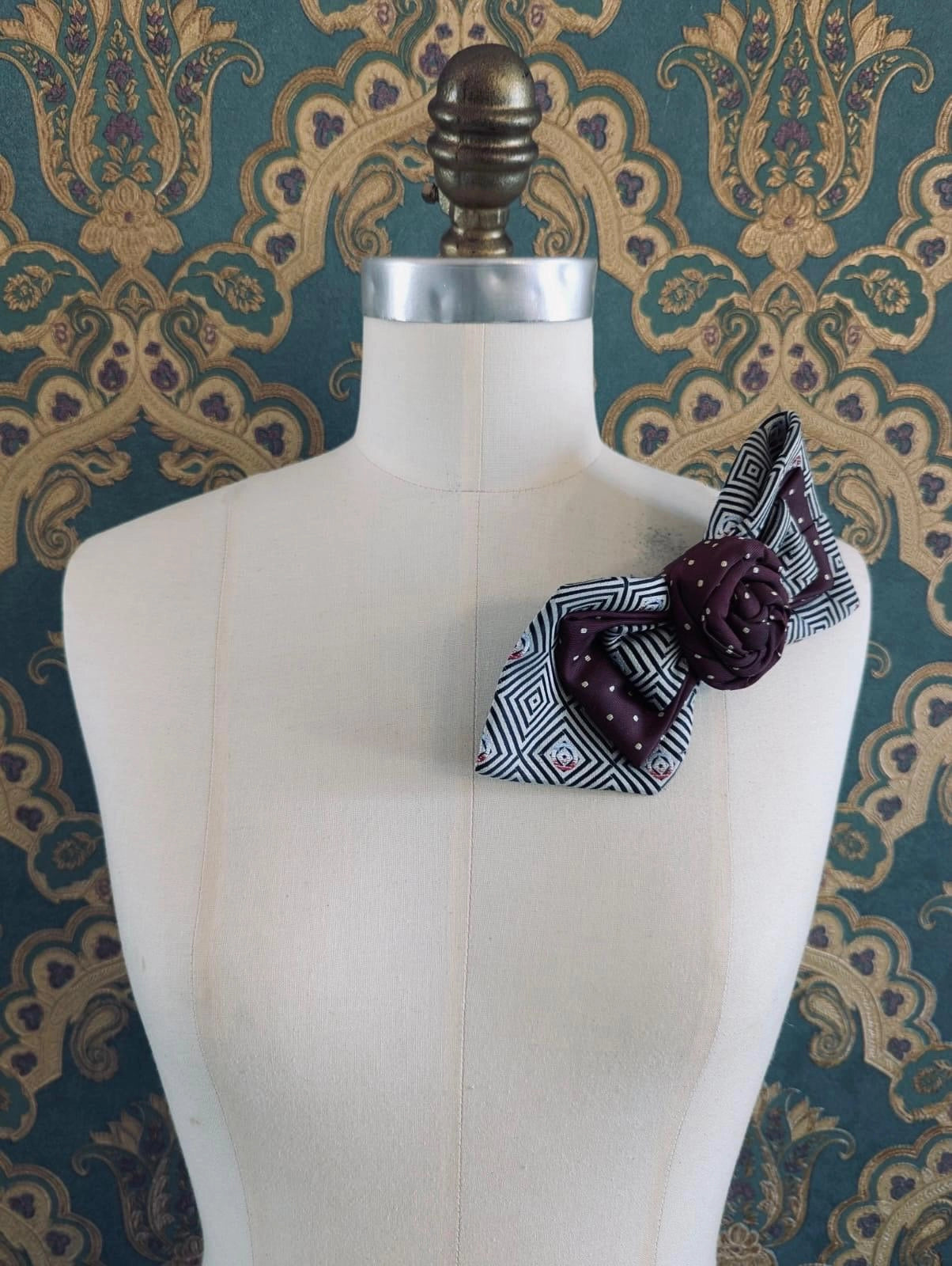 Broochella Bolivar Cravatte hand made up cycled vintage tie brooch in maroon-number 10. it is pinned to a mannequin. The background is green and gold damask wallpaper
