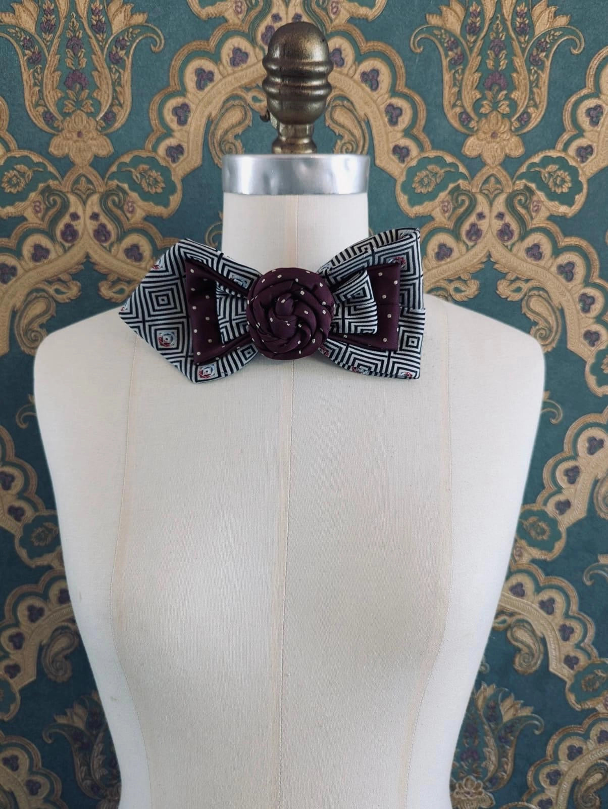 Broochella Bolivar Cravatte hand made up cycled vintage tie brooch in maroon-number 10. it is pinned to a mannequin. The background is green and gold damask wallpaper