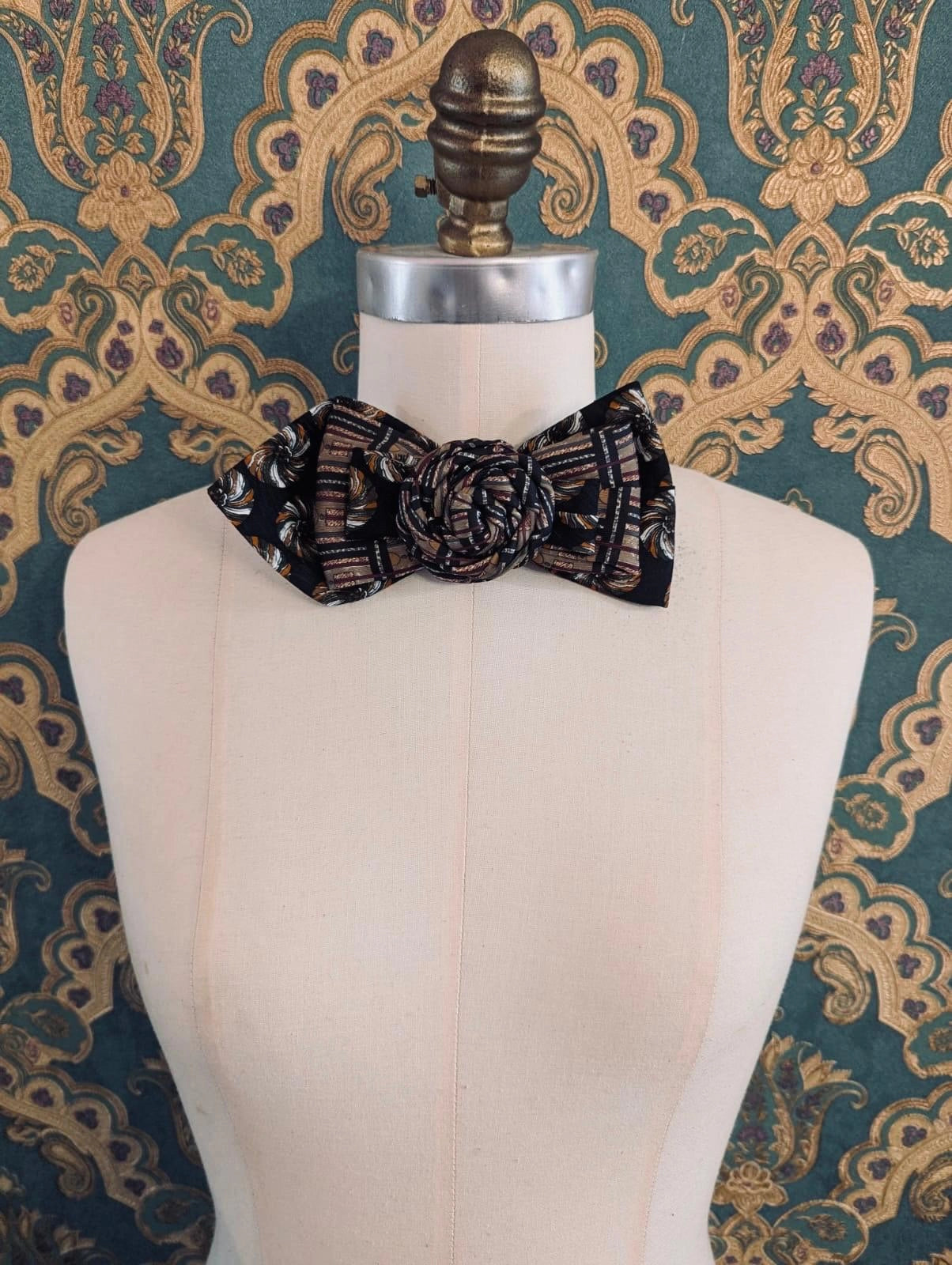 A Broochella Bolivar vintage upcycled necktie styled on a cream fabric dress form mannequin, featuring a black and brown geometric pattern with a central fabric rose embellishment. The background is green and gold damask wallpaper.