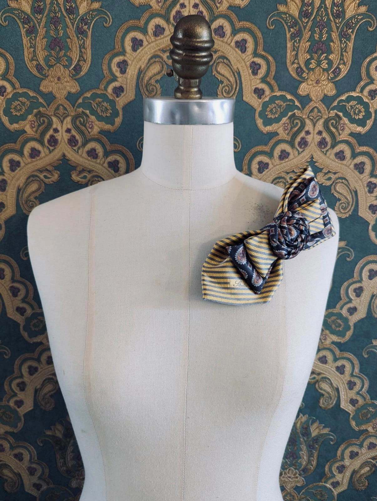 A Broochella Bolivar vintage upcycled necktie styled on a cream fabric dress form mannequin, featuring a yellow and grey geometric pattern with a central fabric rose embellishment. The background is green and gold damask wallpaper.