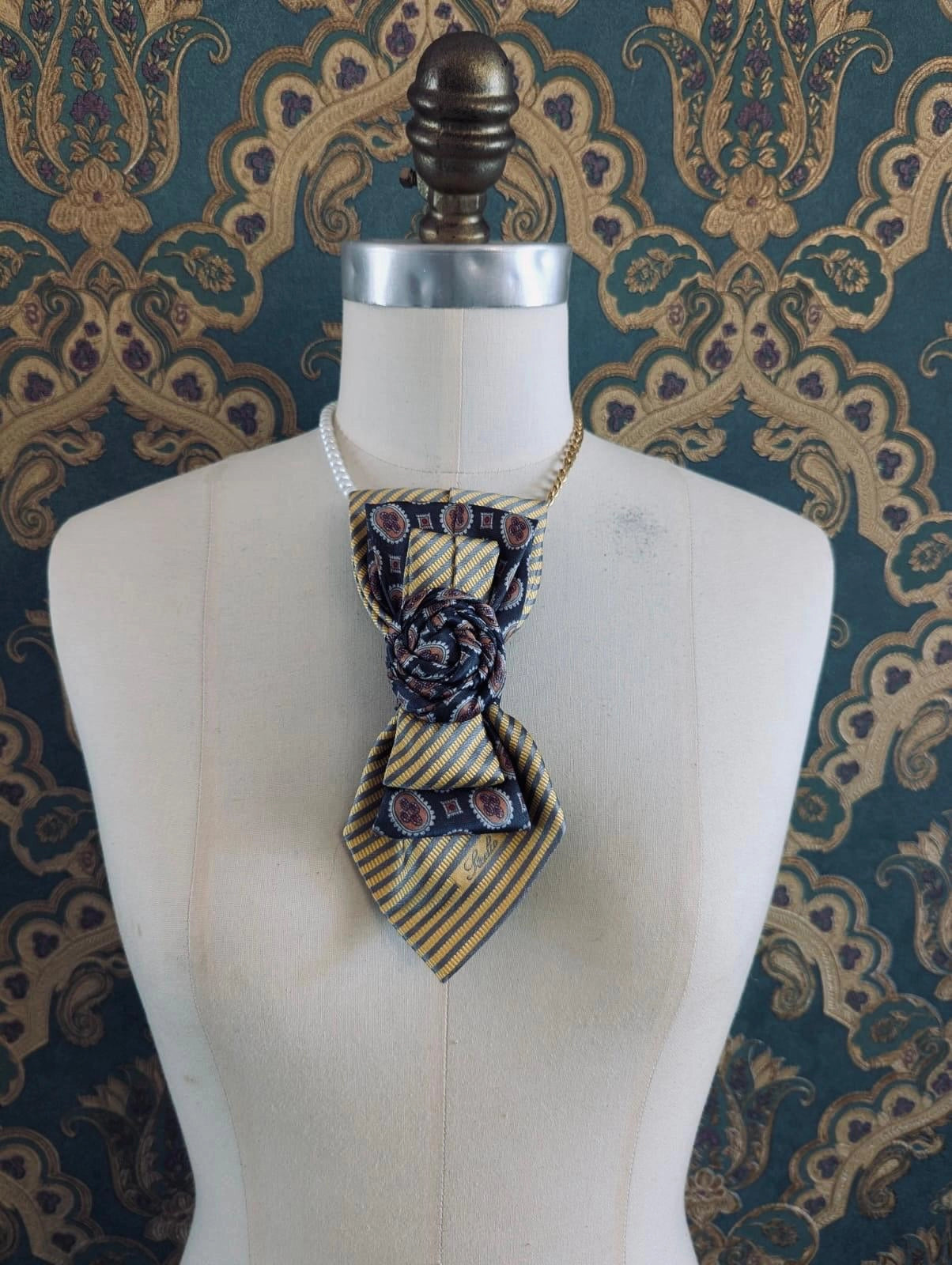 A Broochella Bolivar vintage upcycled necktie styled on a cream fabric dress form mannequin, featuring a yellow and grey geometric pattern with a central fabric rose embellishment and a gold chain strap. The background is green and gold damask wallpaper.