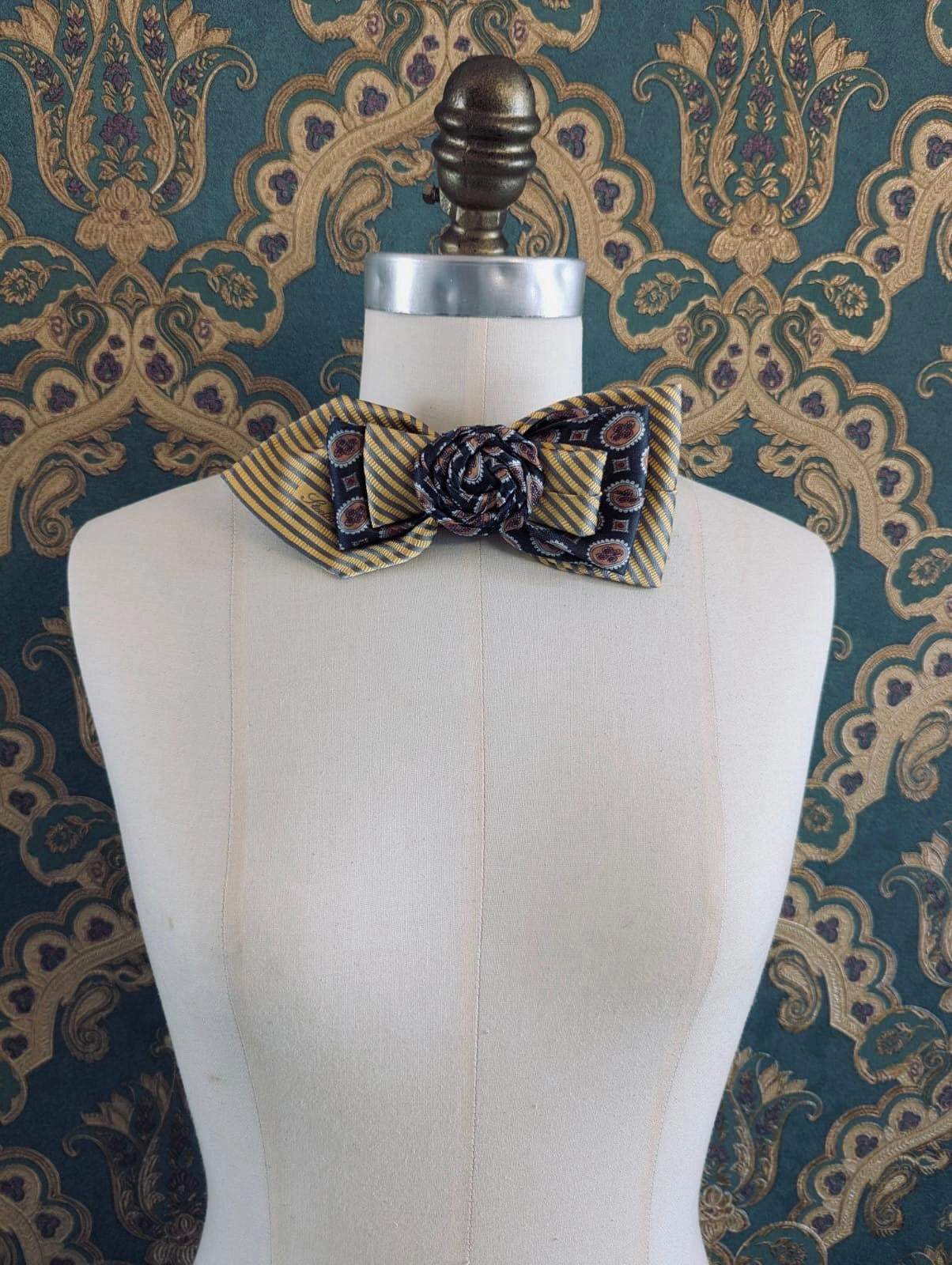 A Broochella Bolivar vintage upcycled necktie styled on a cream fabric dress form mannequin, featuring a yellow and grey geometric pattern with a central fabric rose embellishment. The background is green and gold damask wallpaper.