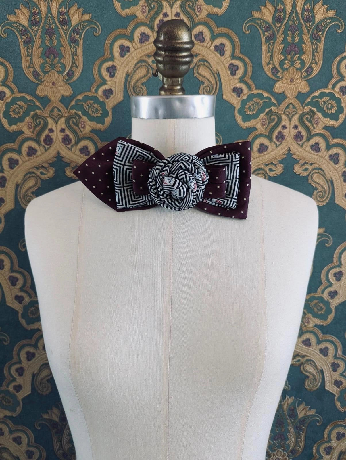 A Broochella Bolivar vintage upcycled necktie styled on a cream fabric dress form mannequin, featuring a burgundy and white geometric pattern with a central fabric rose embellishment. The background is green and gold damask wallpaper.