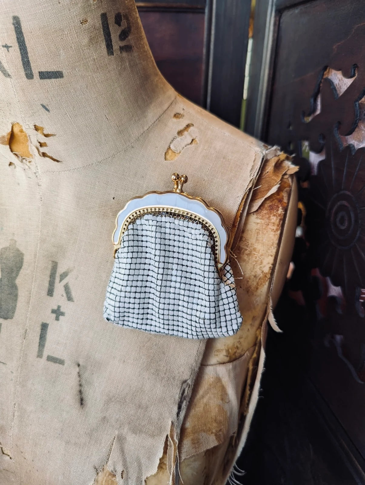 Broochella Borsellino vintage, white, glomesh purse brooch. It has a gold and white metal frame and clasp. It is pinned to a vintage mannequin The background is a decorative, dark wood screen. 