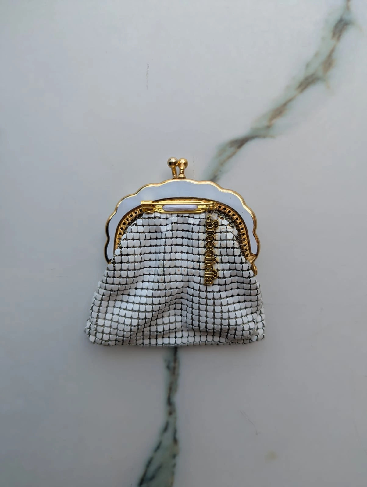 Broochella Borsellino vintage, white, glomesh purse brooch. It has a gold and white metal frame and clasp. The background is white marble. 