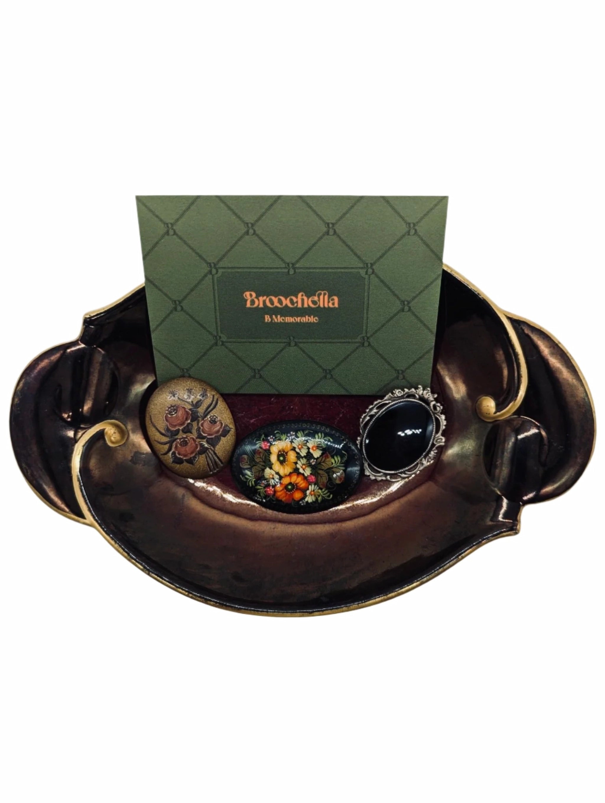 Broochella Botticini brooch trio. There are three brooches. Two are painted with flowers. The other is black with a silver metal frame. They are placed on a wembley ware burgundy and gold tray. There is a broochella business card behind the brooches. The background is white. 