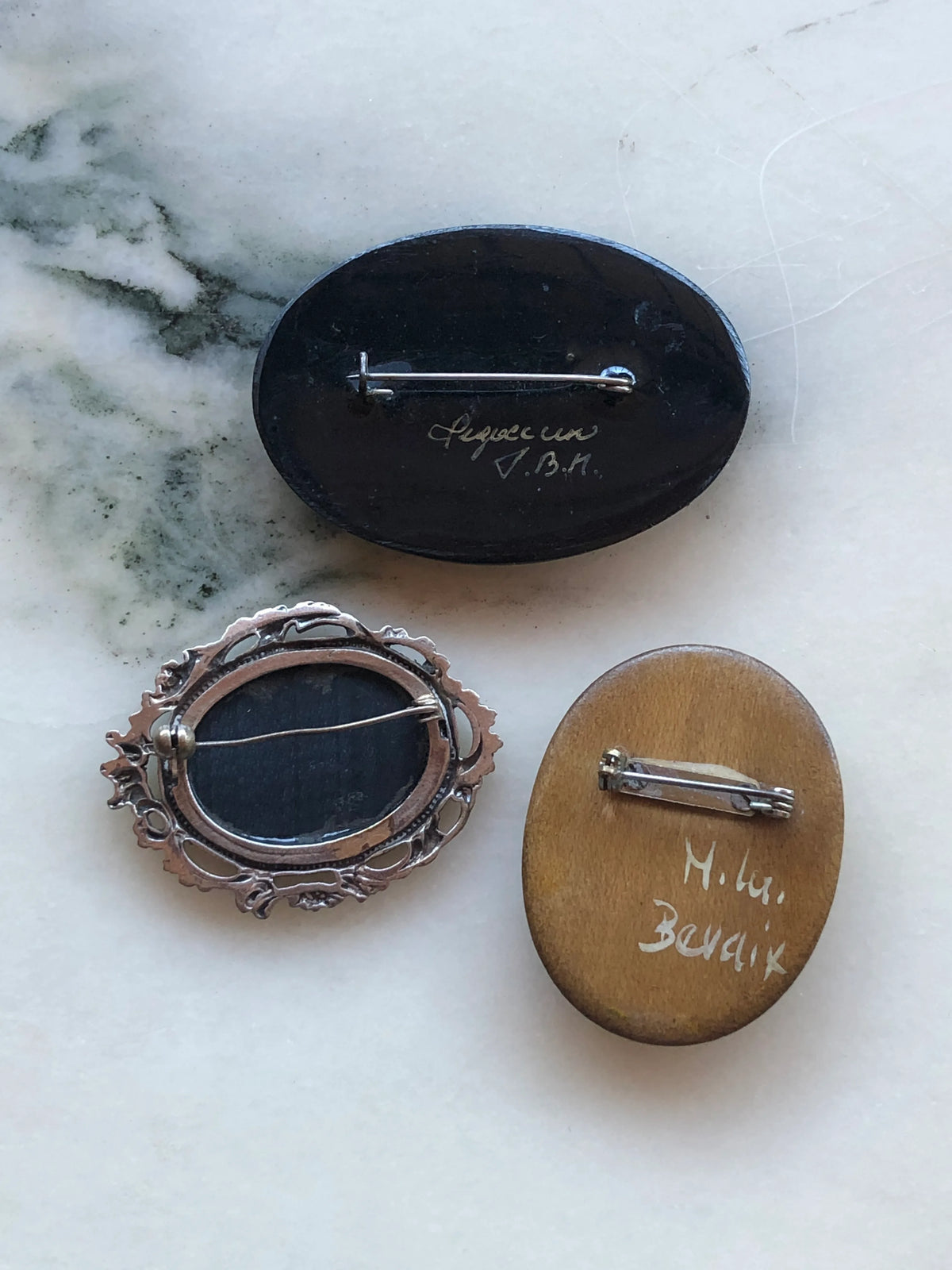 The back view of the Broochella Botticini Vintage Brooch Trio featuring a black hand-painted floral brooch, ornate black oval brooch and hand-painted rose bouquet brooch on a white marble background. 