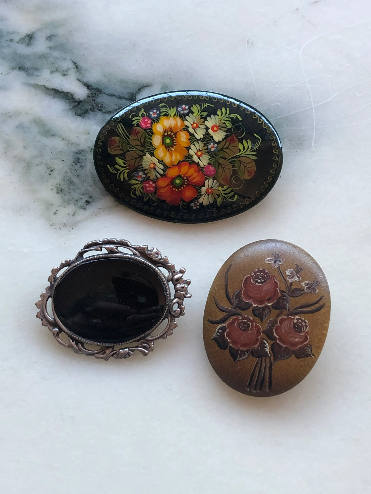 Broochella Botticini Vintage Brooch Trio featuring a black hand-painted floral brooch, ornate black oval brooch and hand-painted rose bouquet brooch on a white marble background.