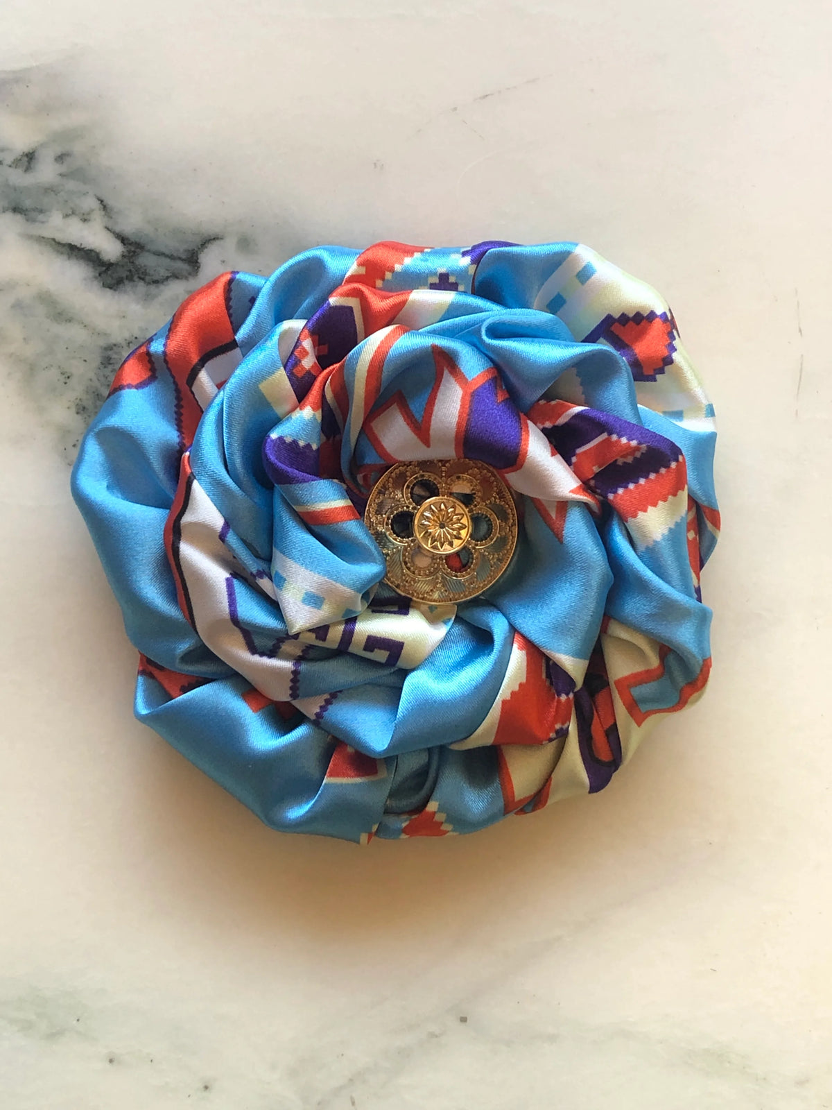 Broochella Men's Bougainville Flower Brooch featuring sculptural blue printed satin petals with red, purple and ivory detailing and an ornate gold-tone centre, photographed on cream marble.