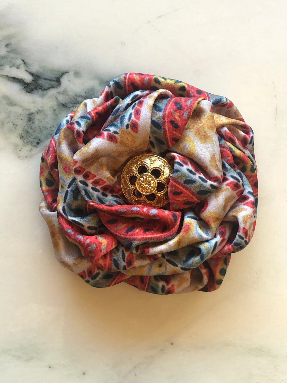 Broochella Men's  Bougainville Flower Brooch featuring sculptural printed satin petals in orange, red, gold and blue tones with an ornate gold-tone centre, photographed on cream marble.