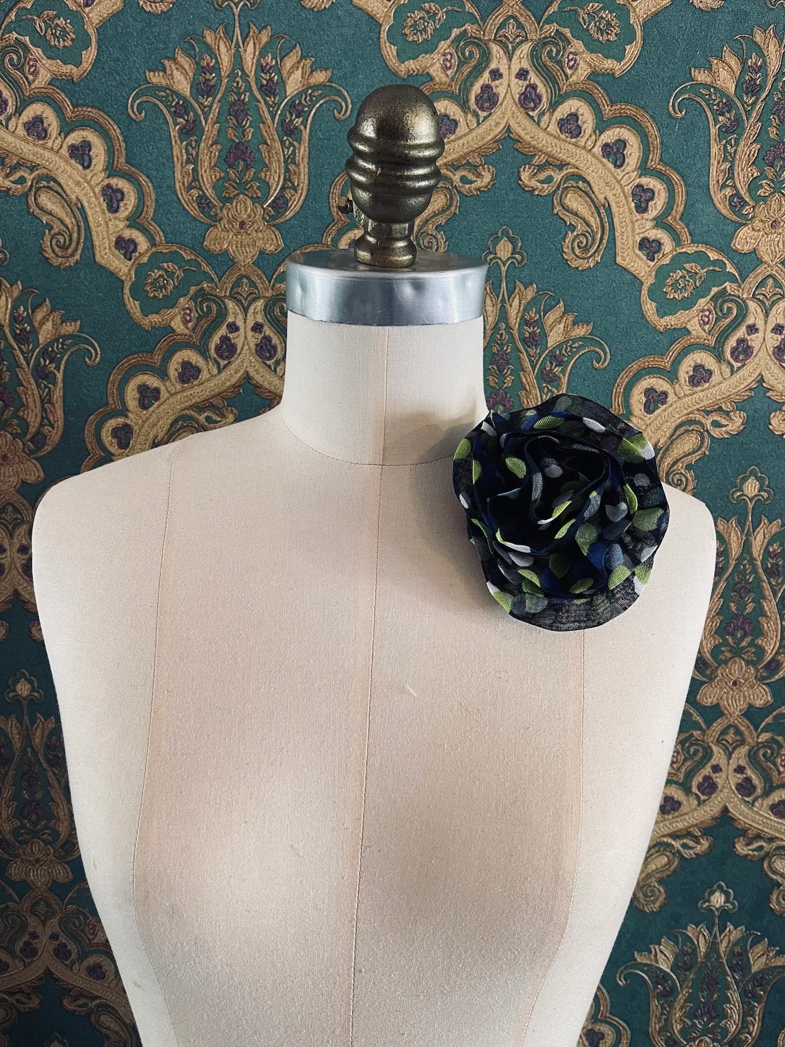 Broochella Brassica handmade fabric flower brooch in navy with blue and green polka dots. Styled on a mannequin. The background is green and gold damask wallpaper