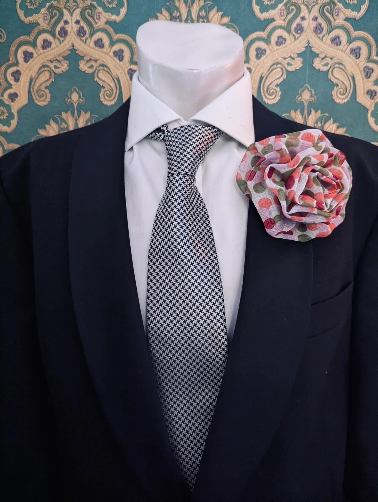 A men's formal outfit displayed on a white mannequin torso, featuring a dark navy suit jacket, a white dress shirt, a gray and white patterned tie, and the Broochella brassica men's flower brooch in green and pink polka dots on the lapel, set against a teal and gold damask wallpaper background.