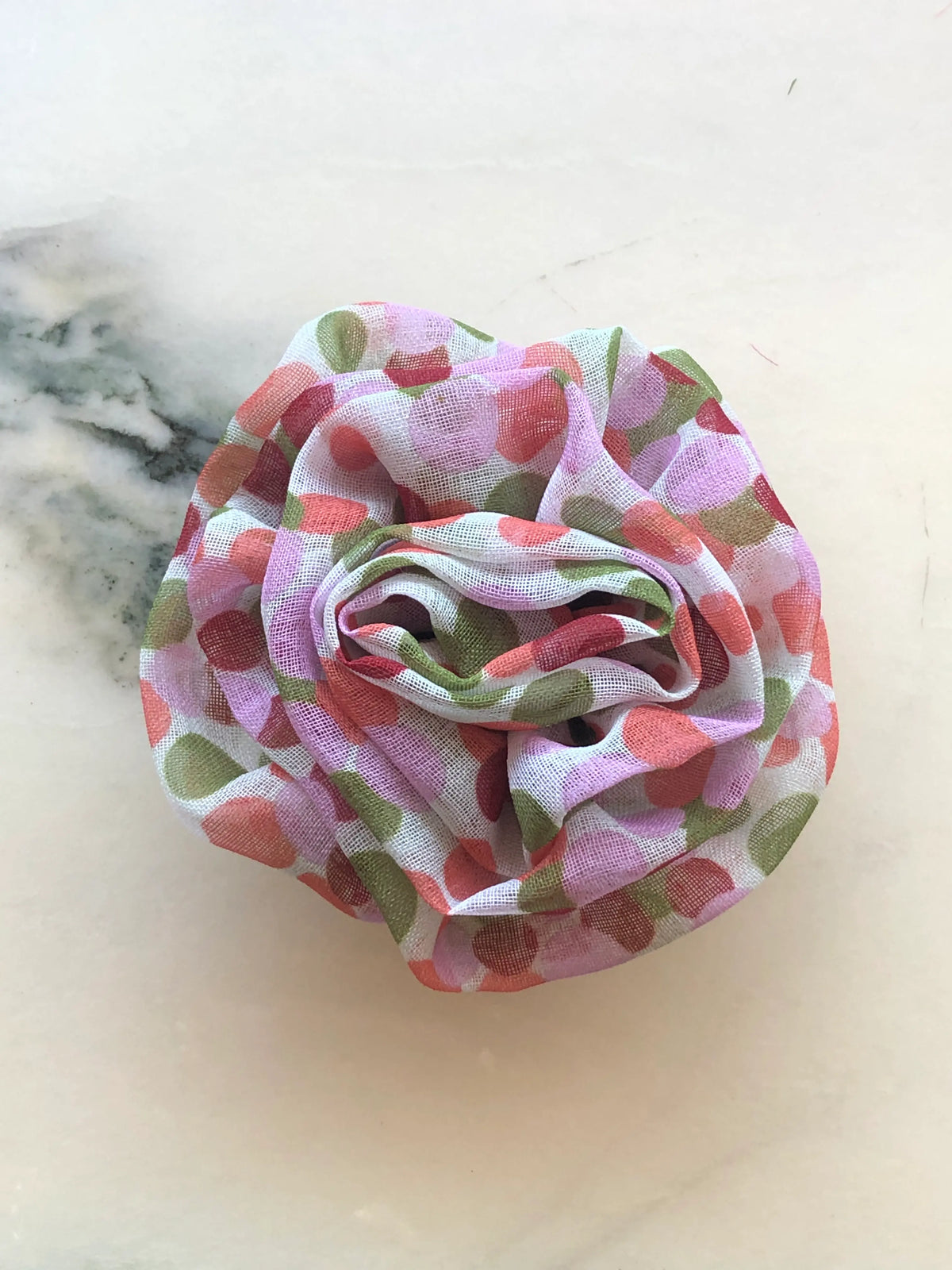 Broochella Brassica Flower Brooch in pink, featuring sculptural sheer fabric petals with a playful print in pink, coral and green, arranged in a full rose-like design on a cream marble background.