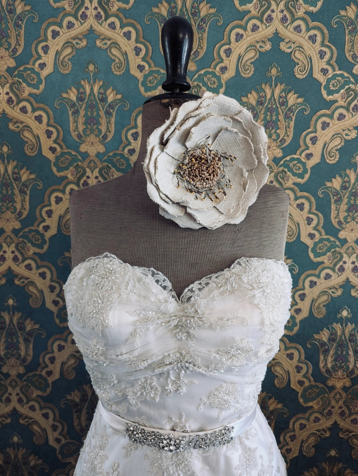 Broochella bridal anemone hand beaded, large, beige, jute wedding flower brooch. Pictured on a mannequin that is wearing a white, beaded strapless wedding dress. The background is green and gold damask wallpaper. 